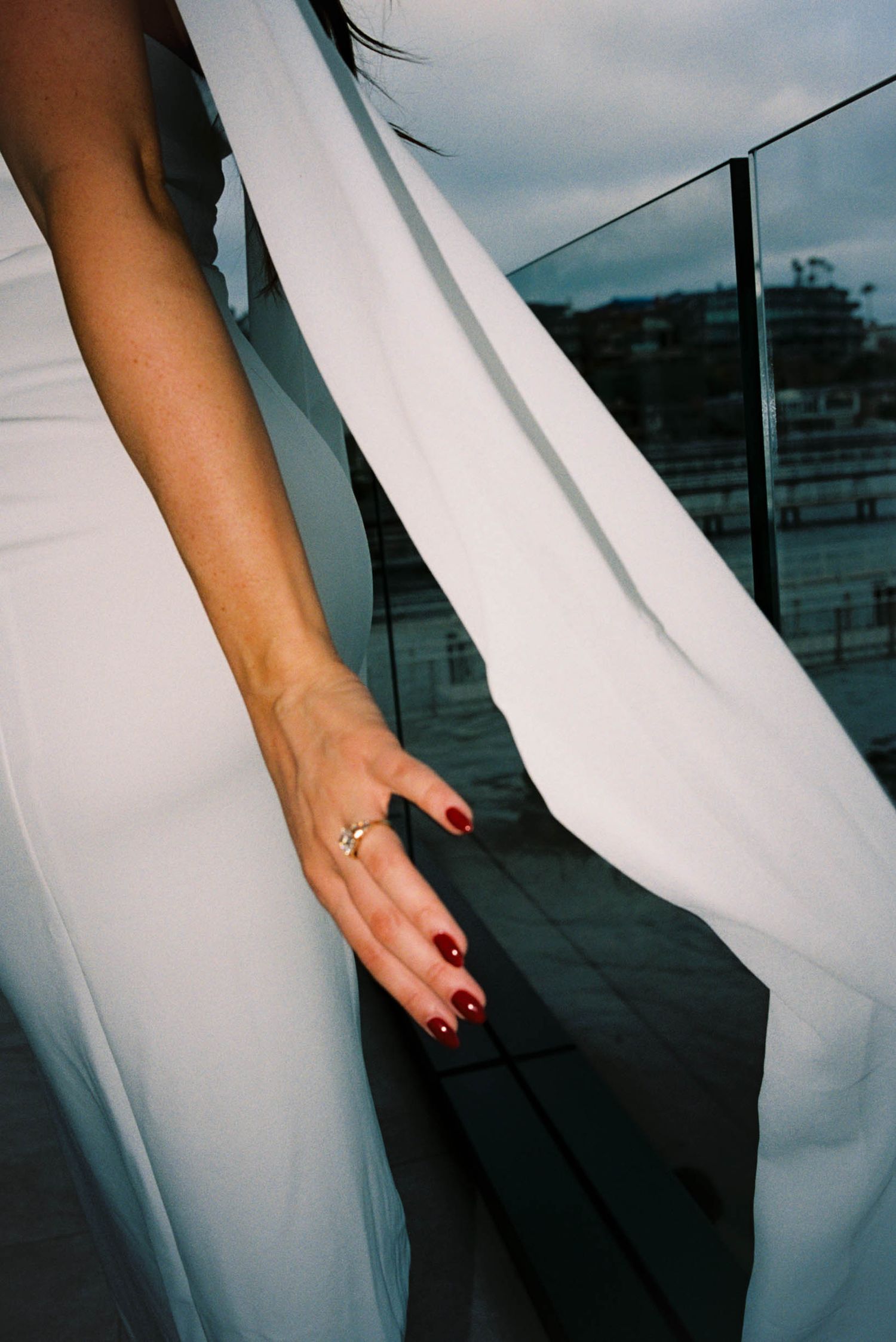 Close-up of white dress fabric and hand with red nail polish wearing engagement ring.