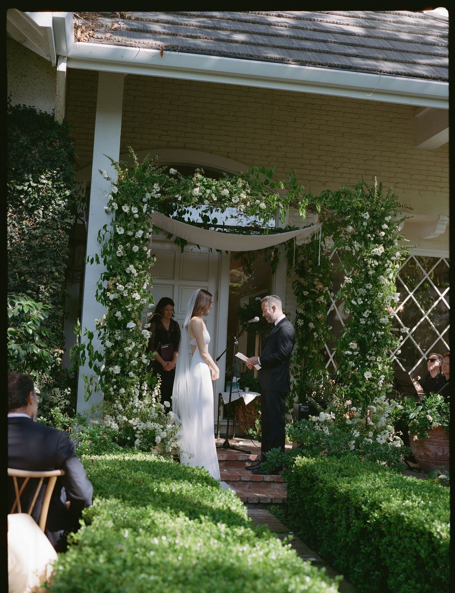 Outdoor wedding ceremony under a lush floral arch with bride, groom, and officiant in a garden setting.
