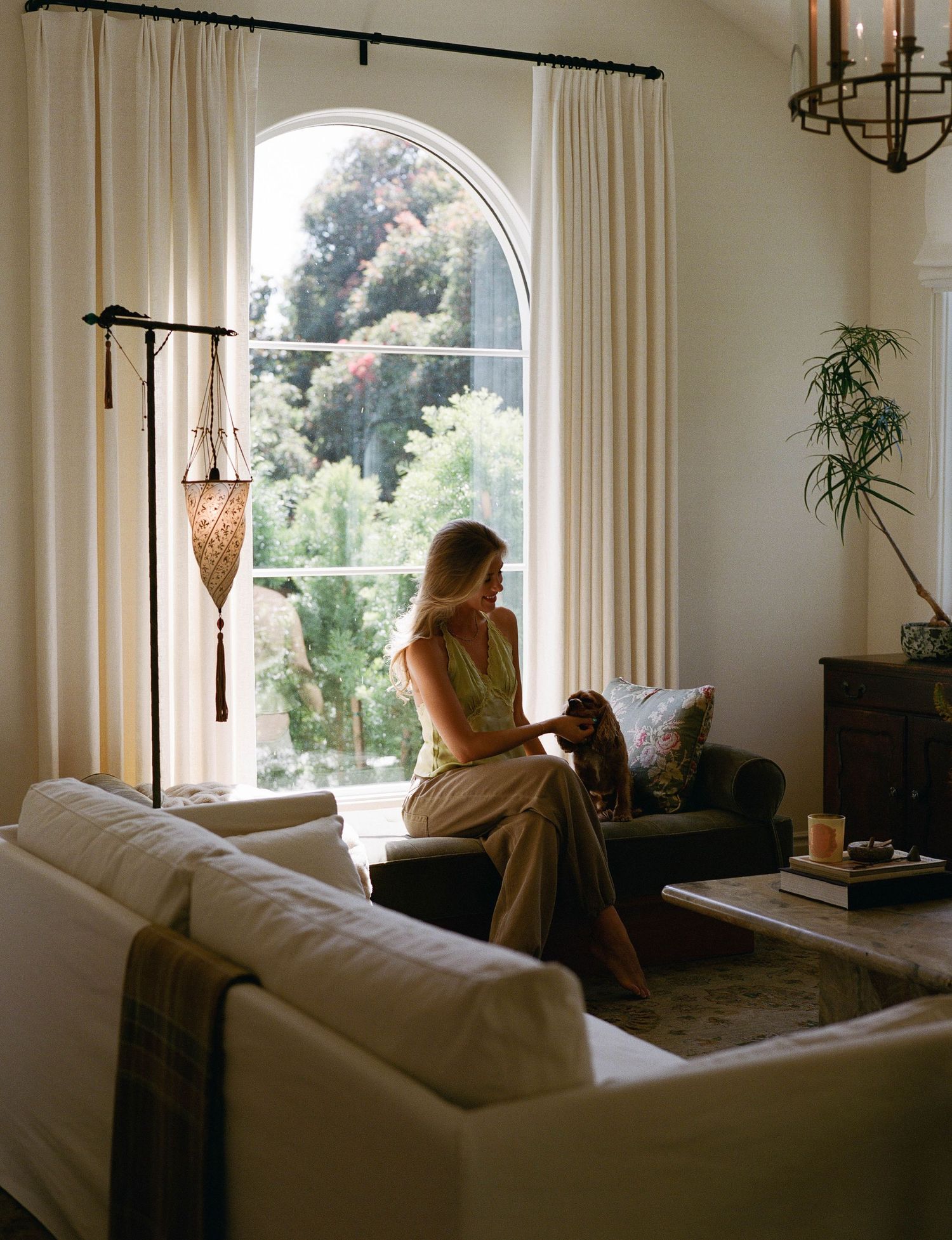 Woman sitting in elegant living room with arched window, white sofa, and lush garden view.
