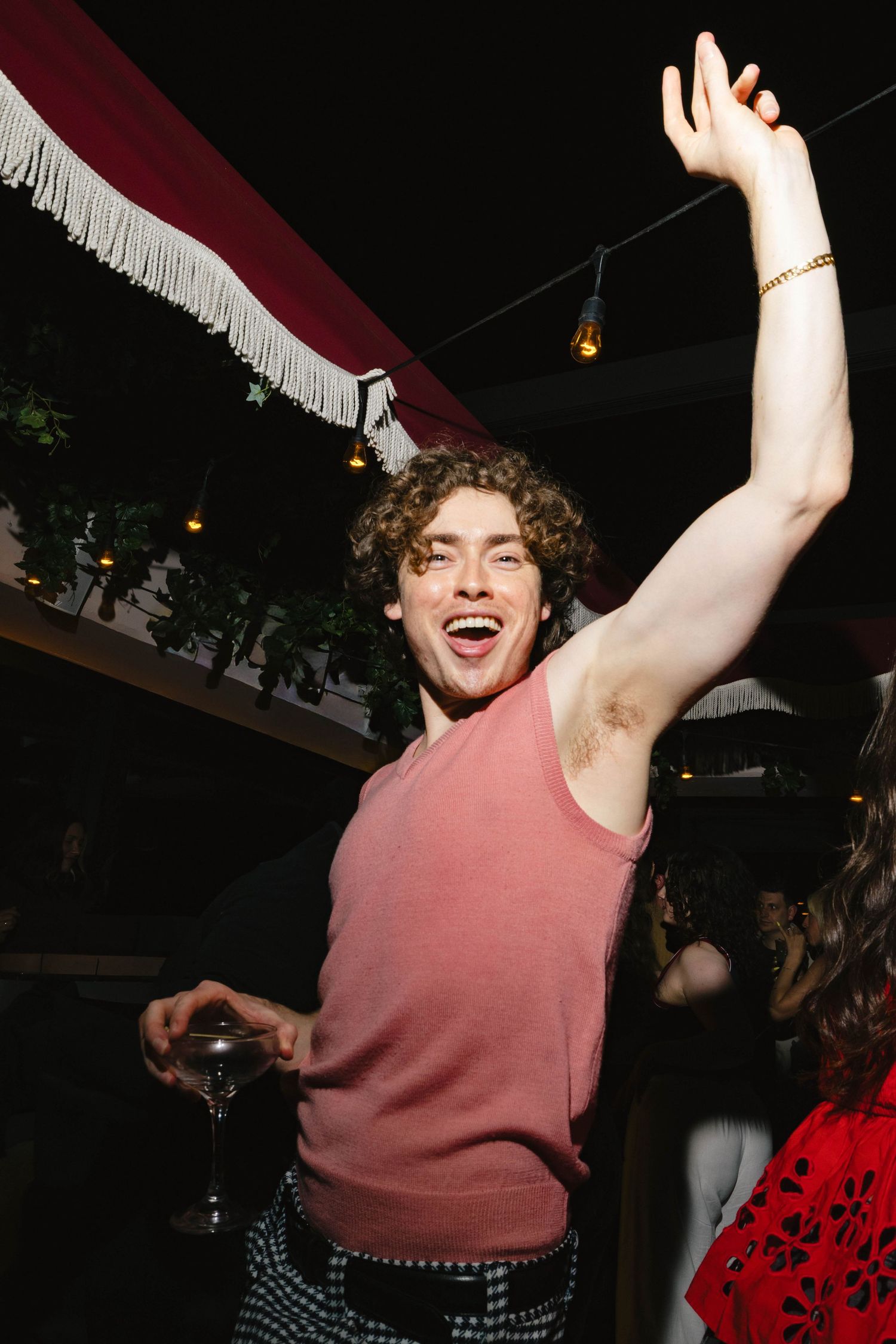 Young man with curly hair dancing at a party, raising his arm while holding a wine glass, wearing a pink tank top.