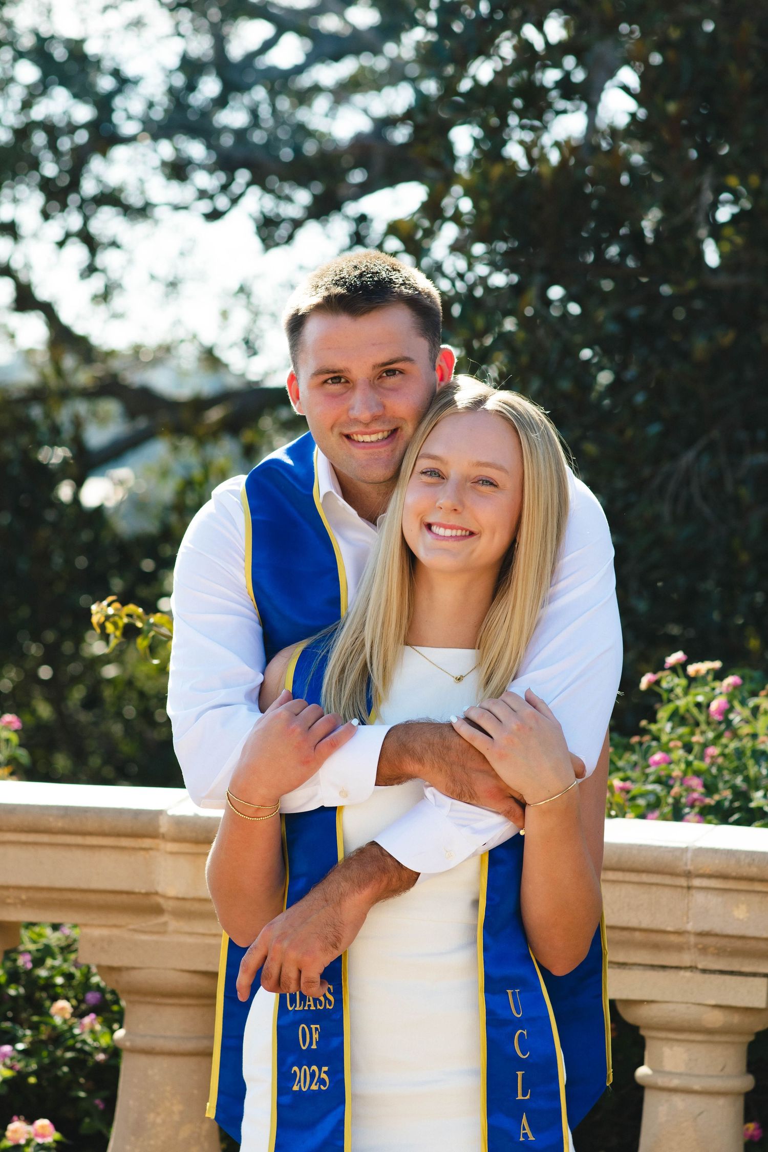Graduates in white outfits with blue sashes embrace while celebrating on campus.
