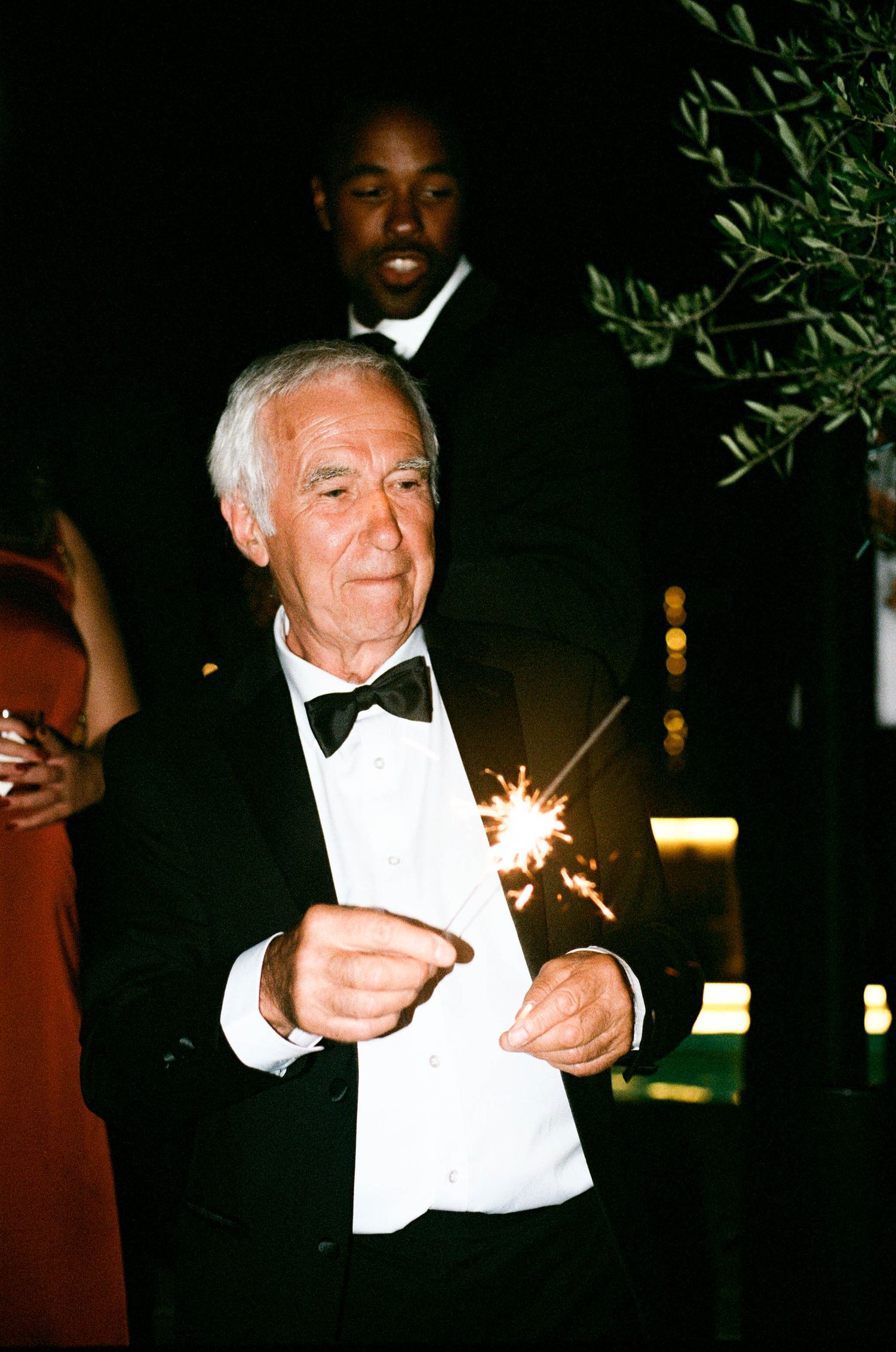 An elderly gentleman in a tuxedo lights sparklers at a nighttime celebration.