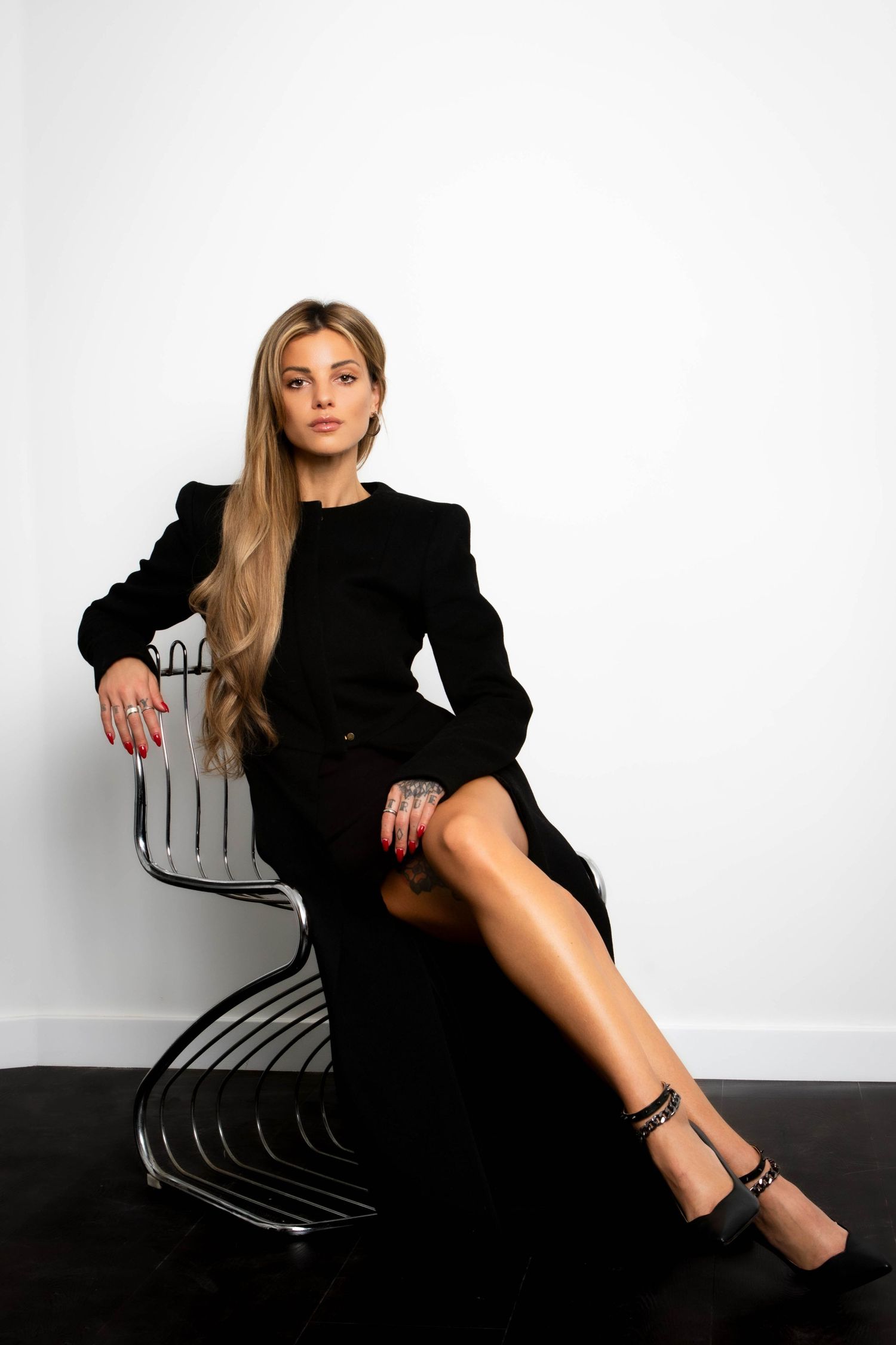 A person in a black dress poses elegantly on a modern curved chair against a white backdrop in a fashion photoshoot series.