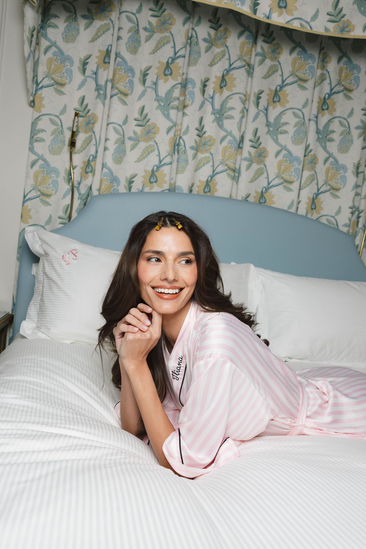 Smiling woman in a pink striped satin pajama robe lies on a white bed with a blue headboard and floral canopy curtains.