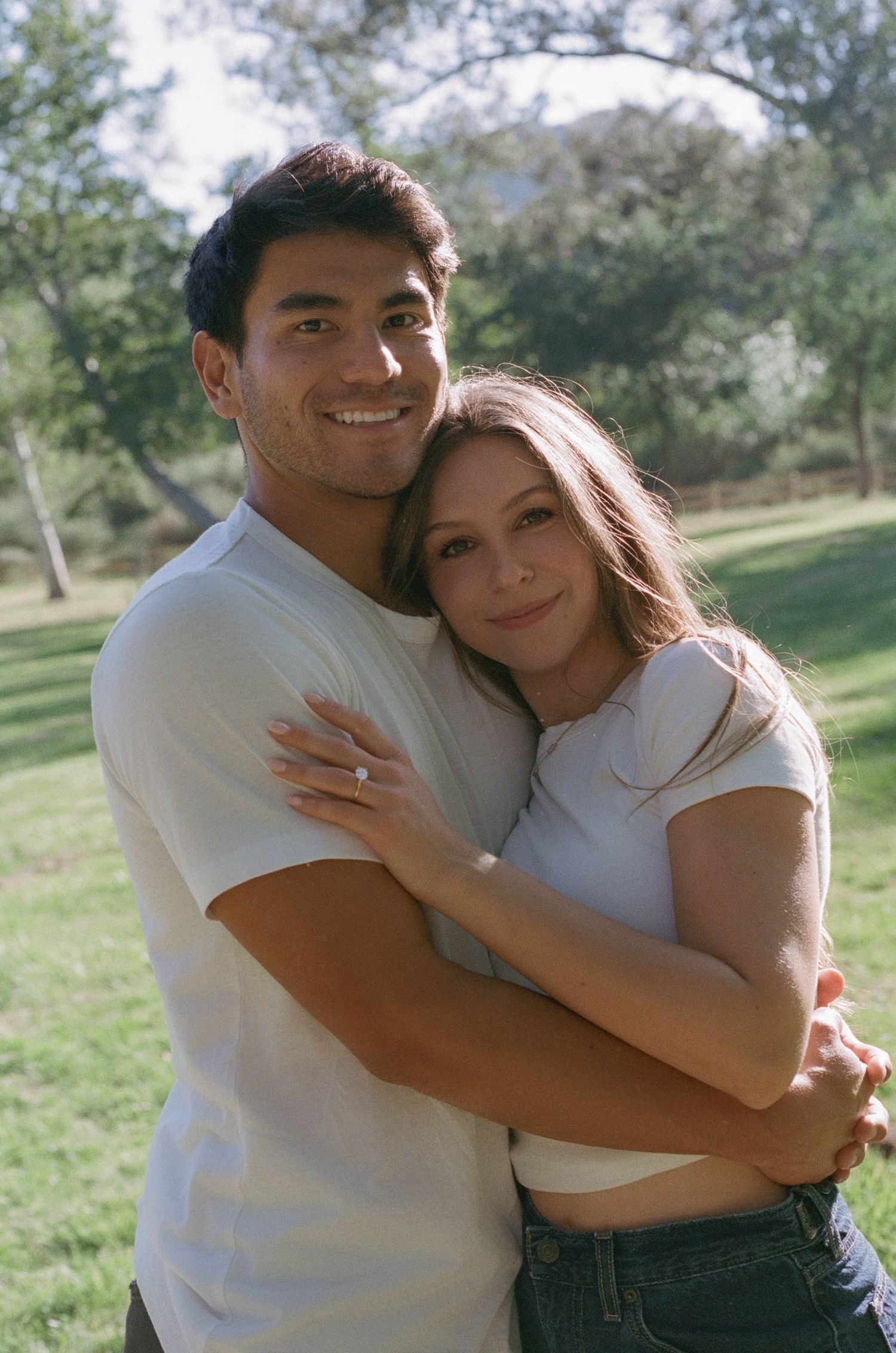 Happy couple embracing outdoors in a park, wearing white shirts, woman showing engagement ring.