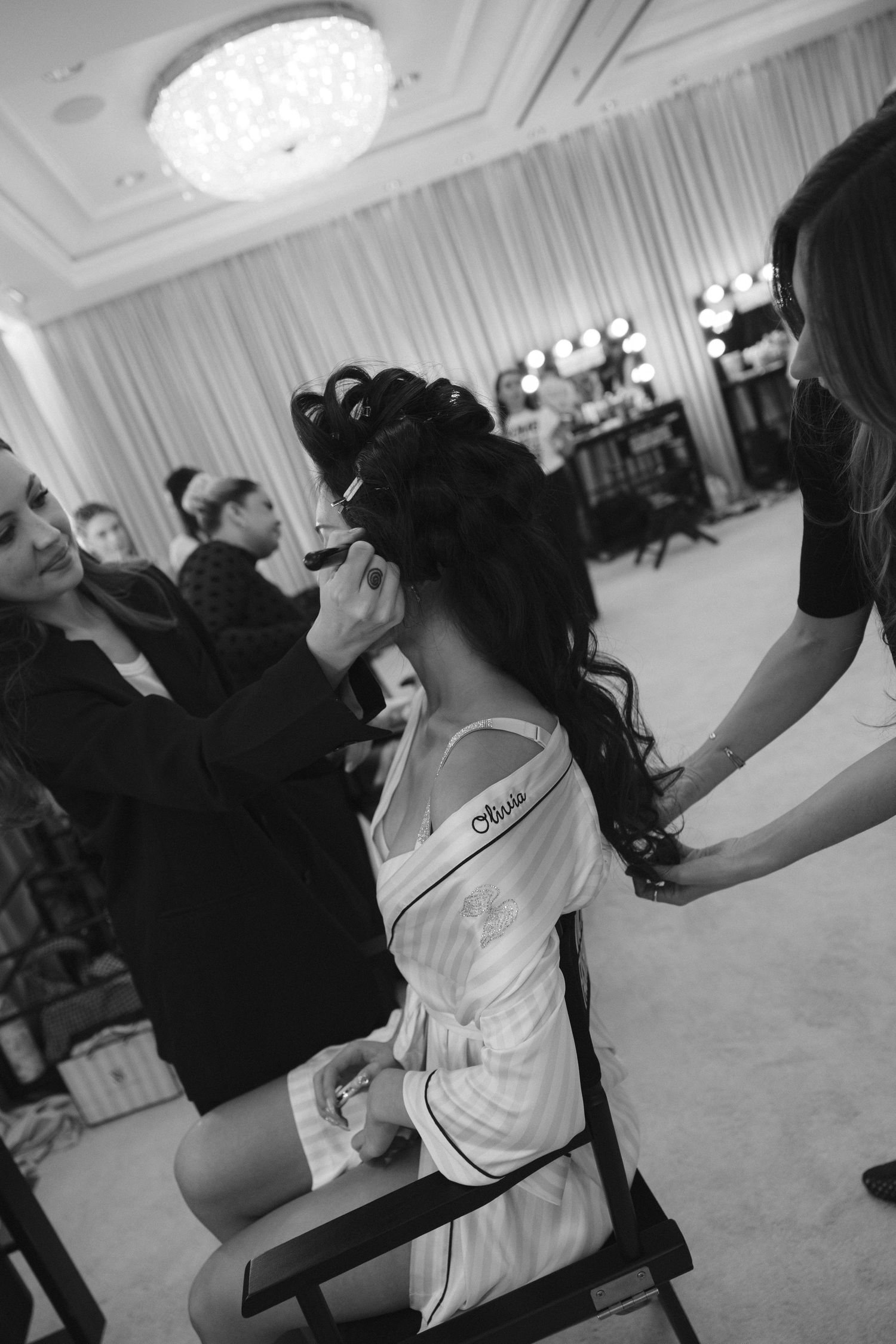 A bride in a monogrammed satin robe sits in a chair while makeup artists style her curled updo in a bright prep room.