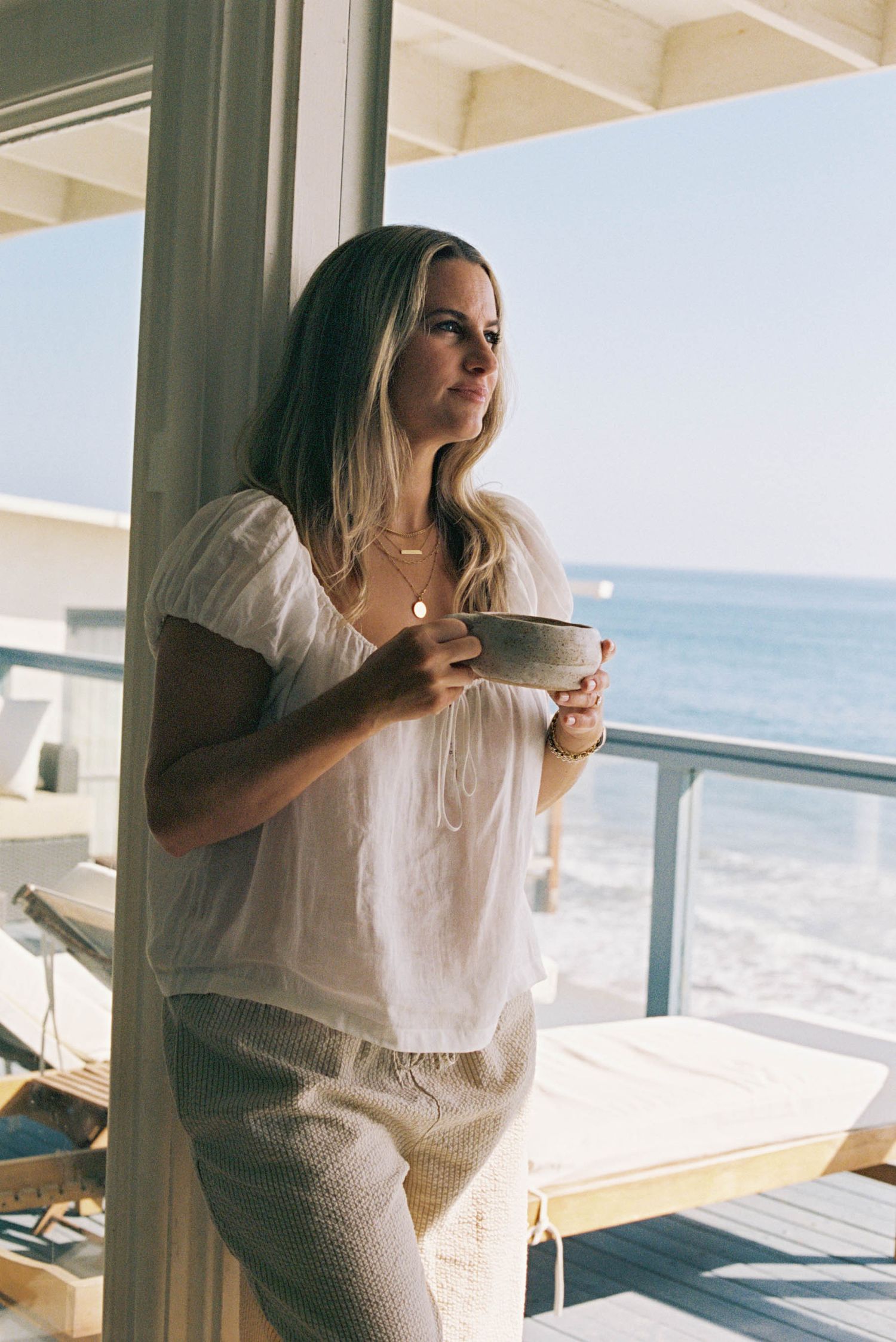 Woman in white top enjoying coffee while looking out at ocean view from bright coastal room with large windows.