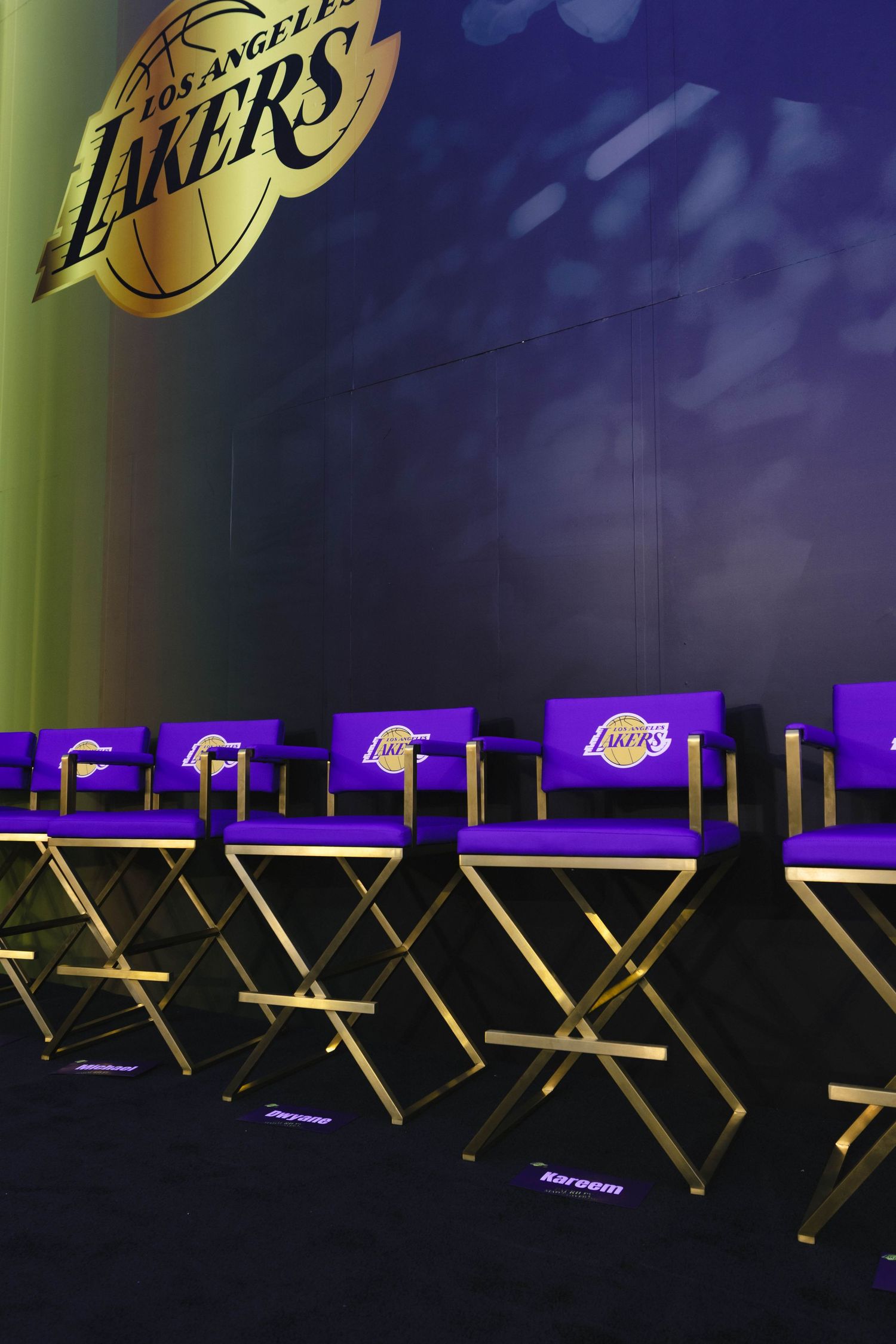 Purple and gold Los Angeles Lakers branded director chairs arranged in a row against a dark Lakers logo backdrop.