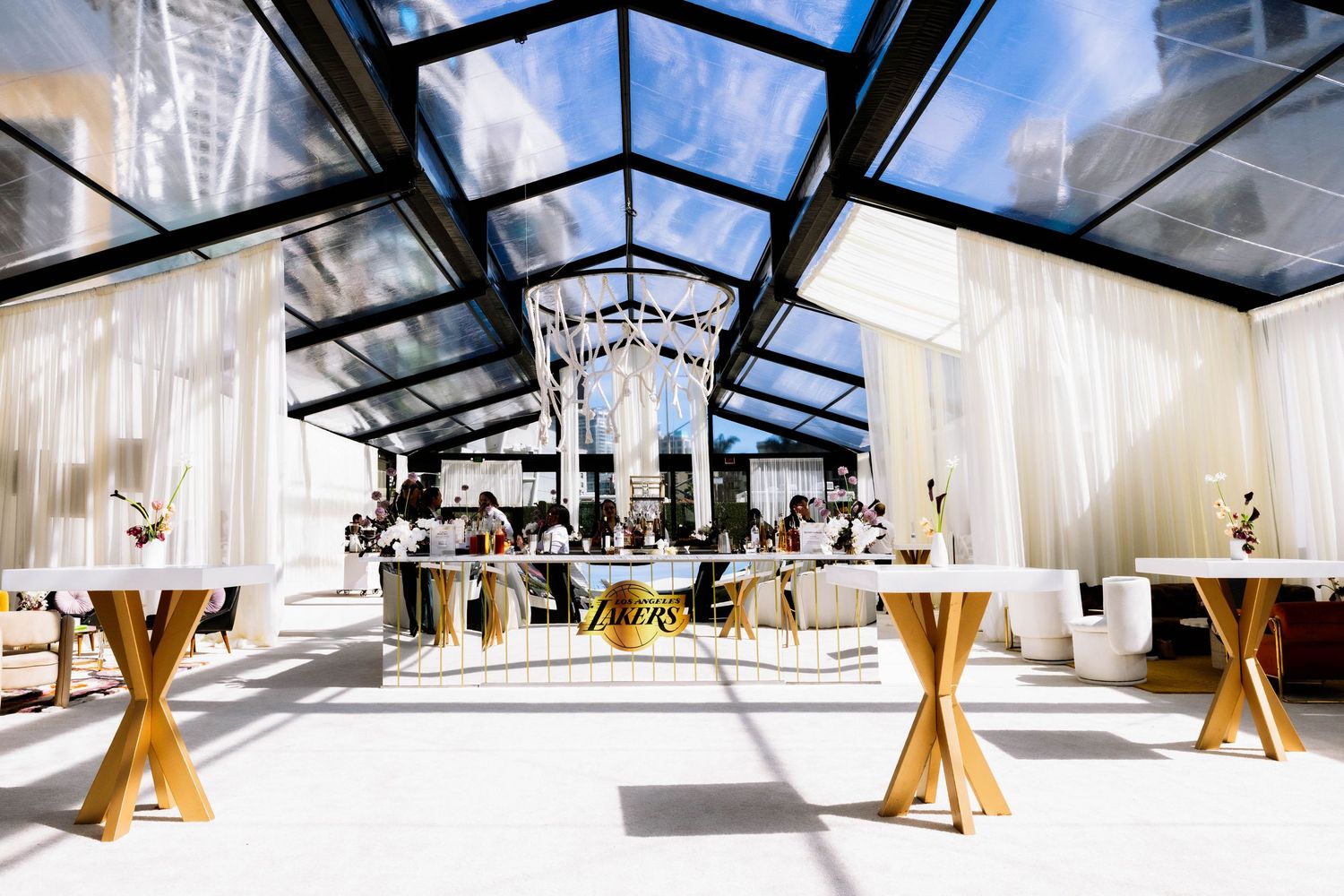 Elegant glass-roofed wedding reception venue with gold accent tables, white draping, and crystal chandelier.