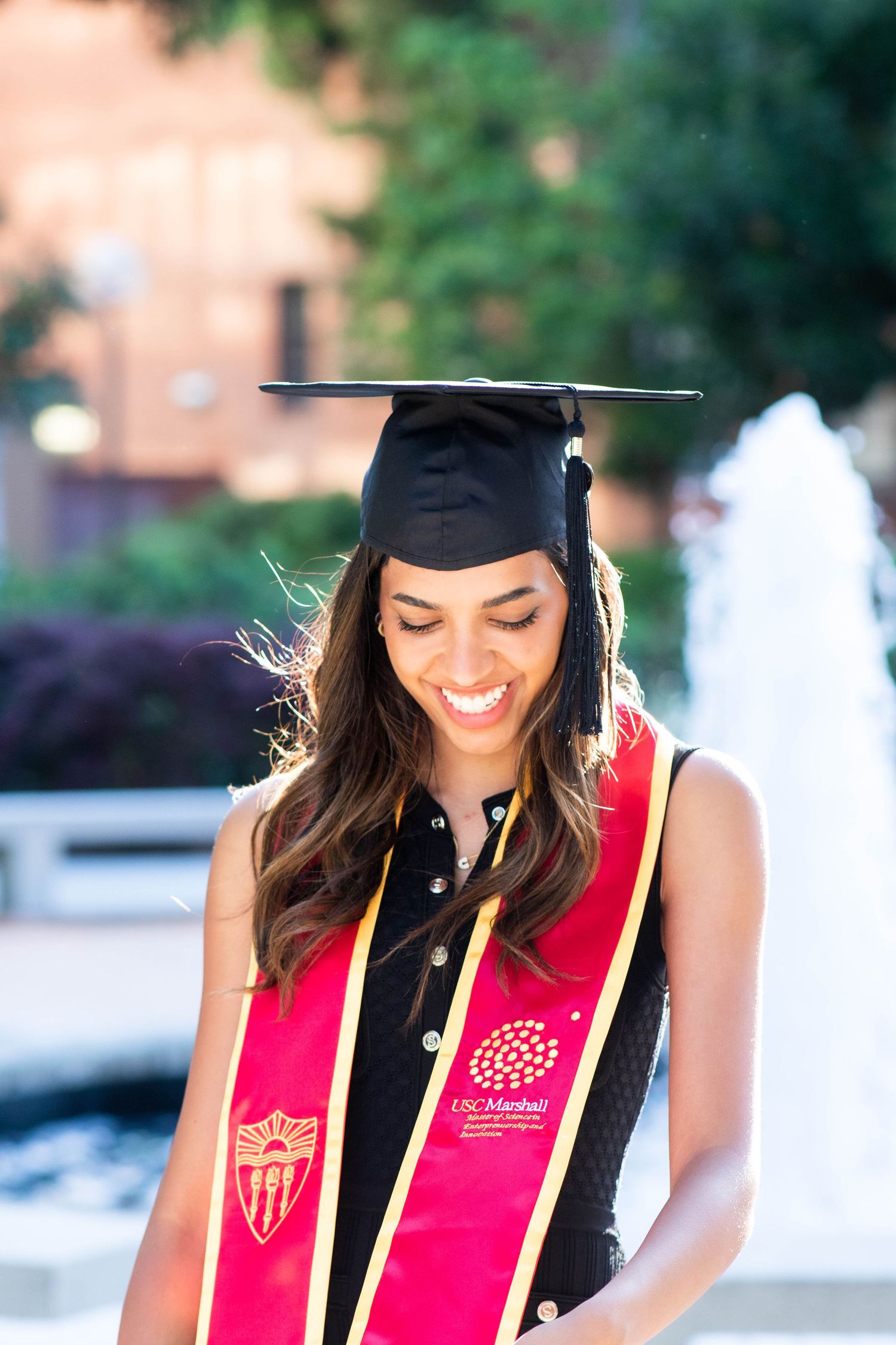 Graduate wearing cap and red graduation stole smiles outdoors on campus.