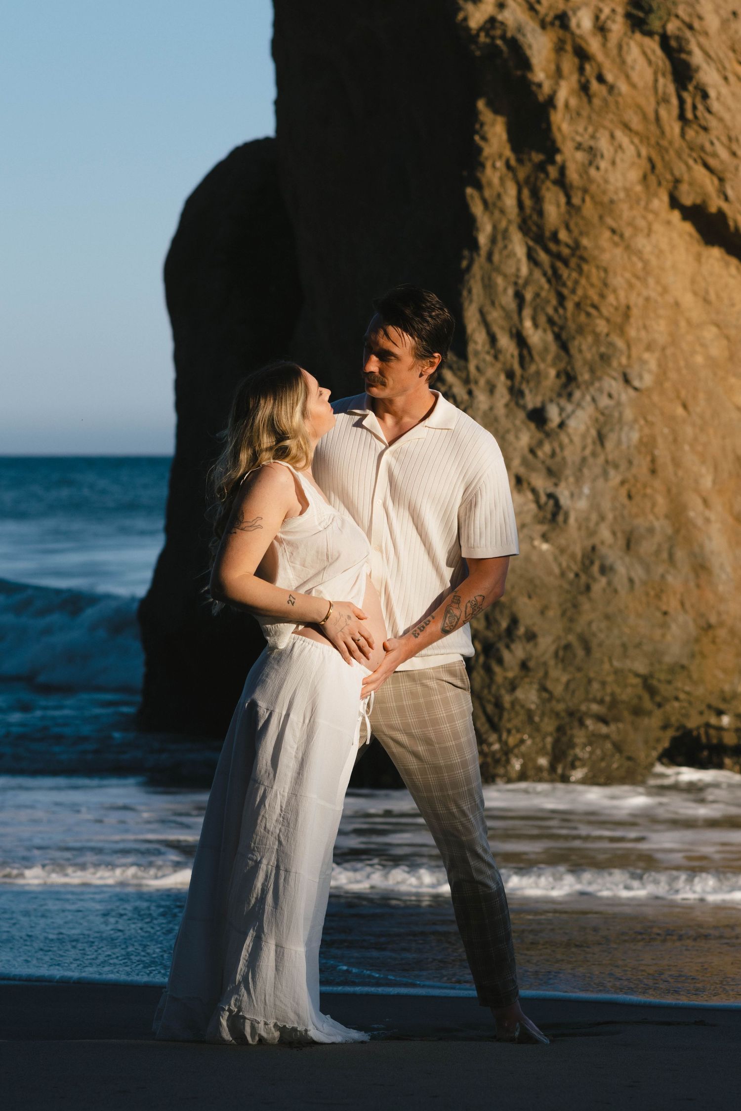 Romantic beach photoshoot with couple dancing near ocean rocks at sunset.