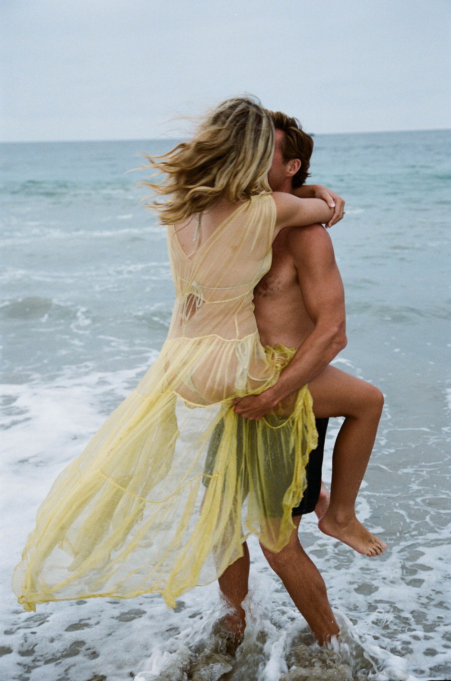 A person in a flowing yellow dress dances and splashes in ocean waves at the beach.