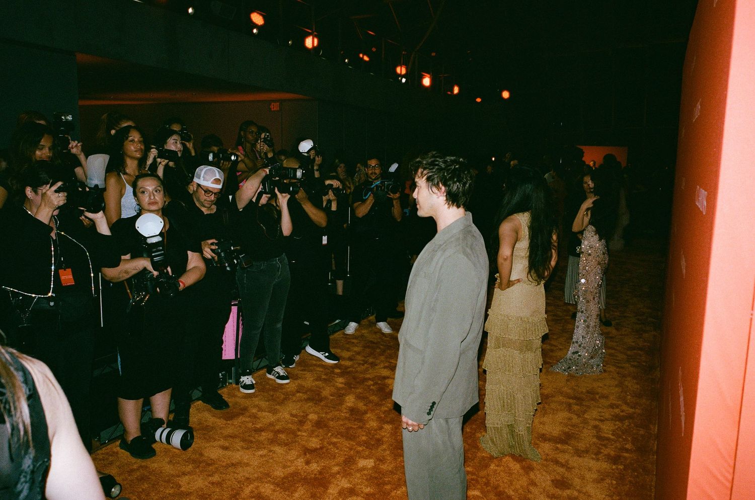 A man in a gray suit stands on a dance floor surrounded by photographers and guests at an indoor event.
