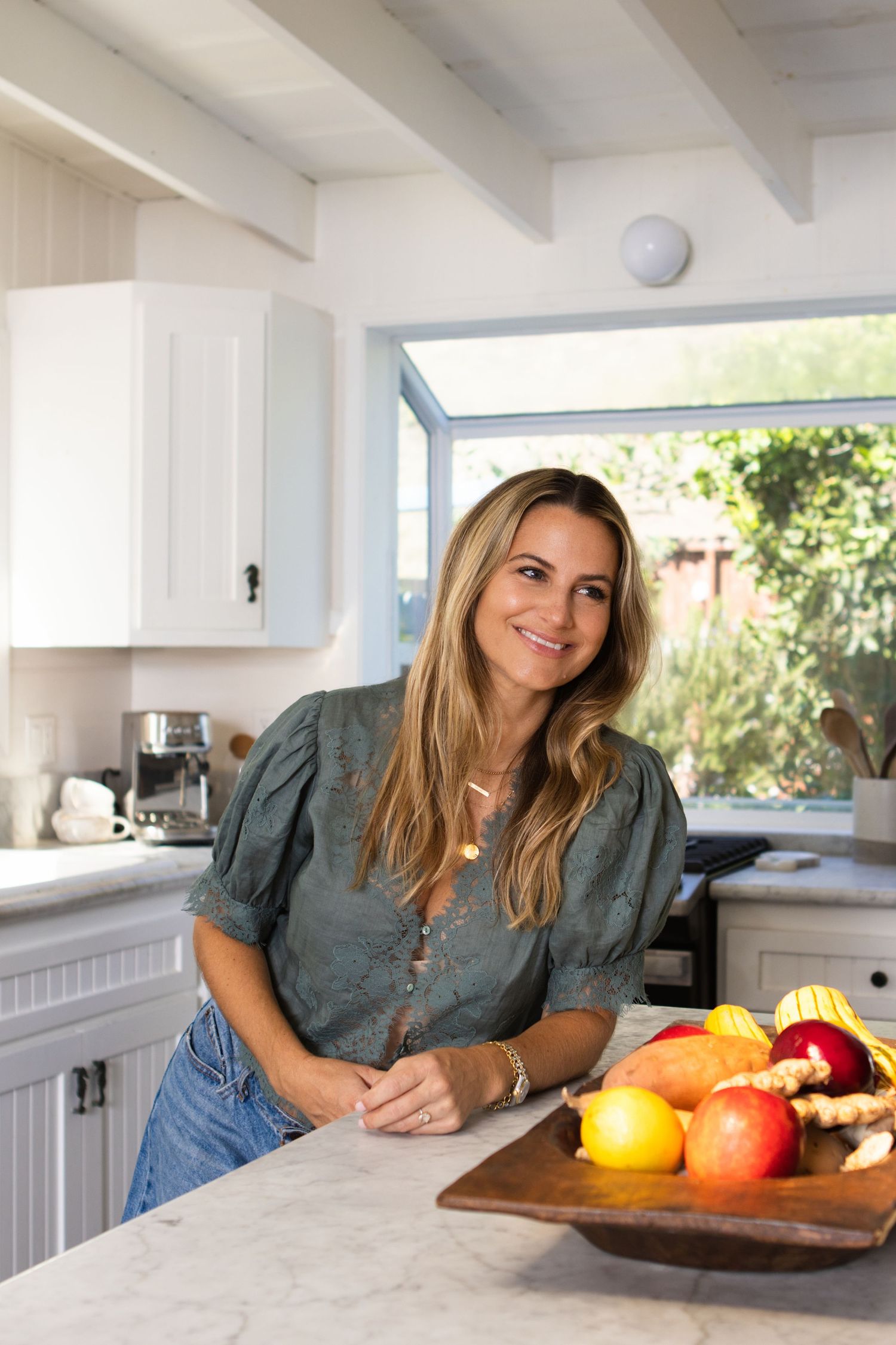 A person in a gray shirt leans on a white kitchen counter near a wooden cutting board with colorful fresh fruit.