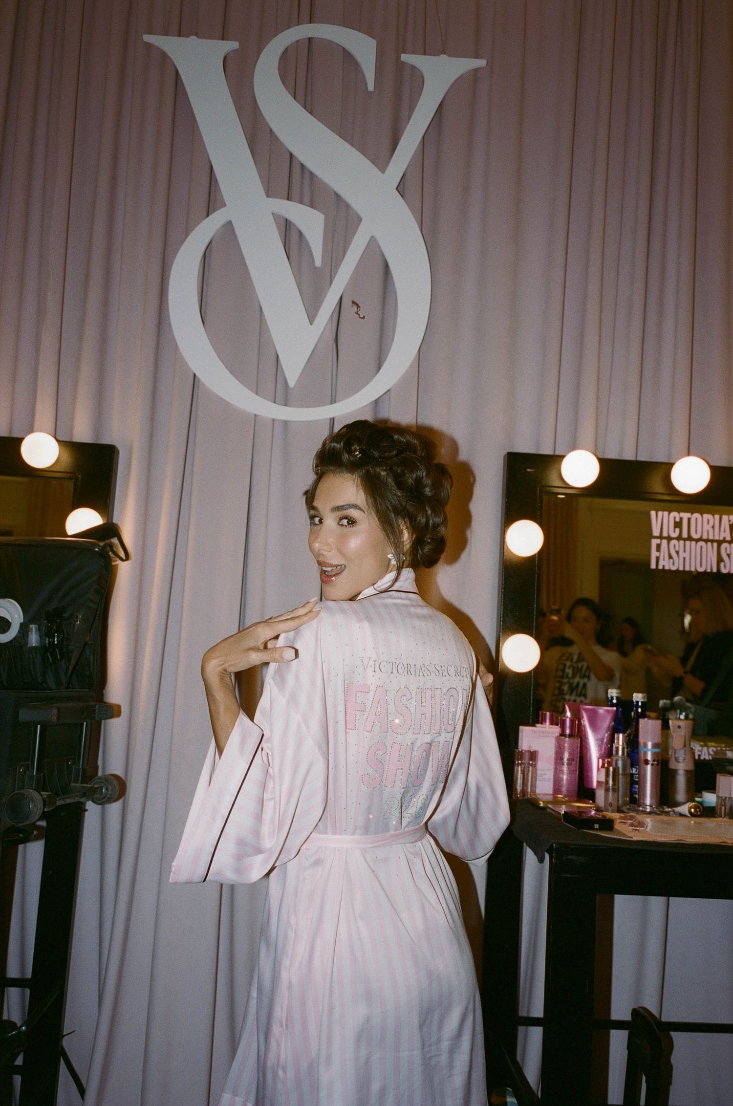 Woman in a pink striped satin robe smiles over her shoulder backstage beside lit vanity mirrors and a Victoria's Secret Fashion Show logo.