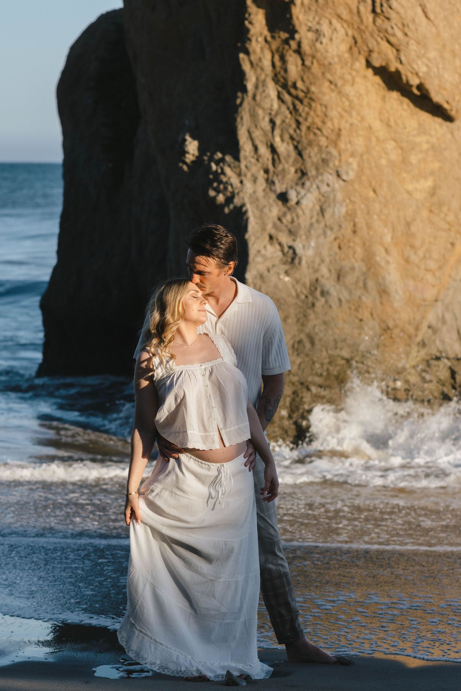 A romantic couple embraces on a beach near rocky cliffs as ocean waves crash behind them.