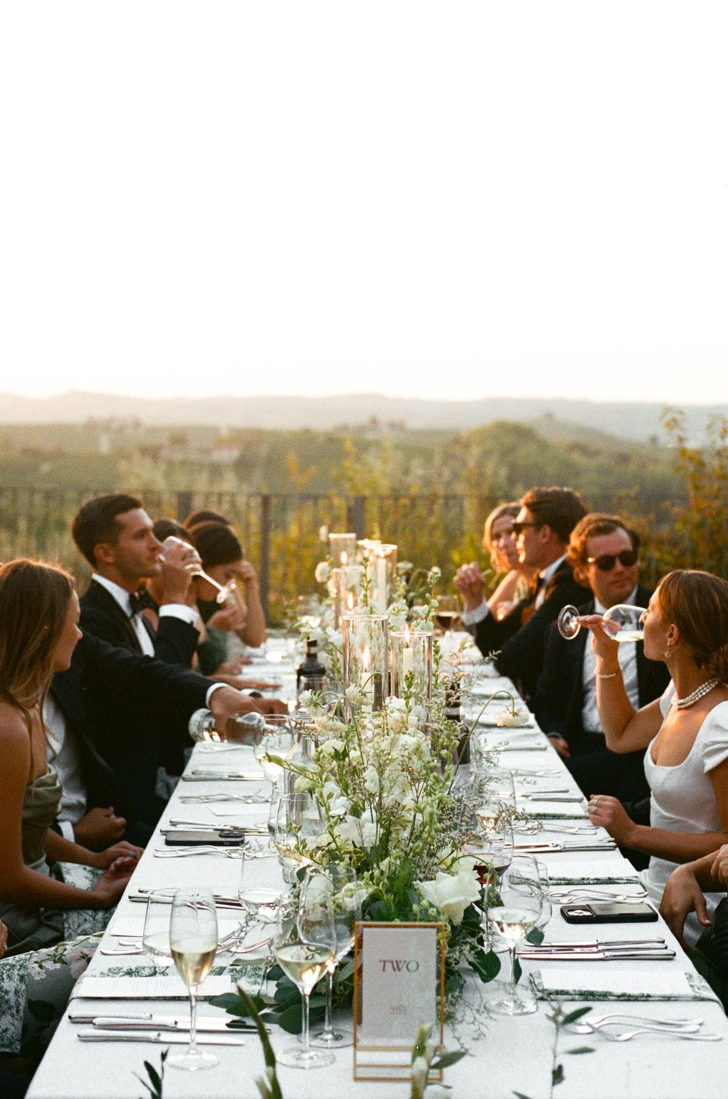 Elegant outdoor wedding reception table setting with white linens and delicate floral centerpieces at sunset.