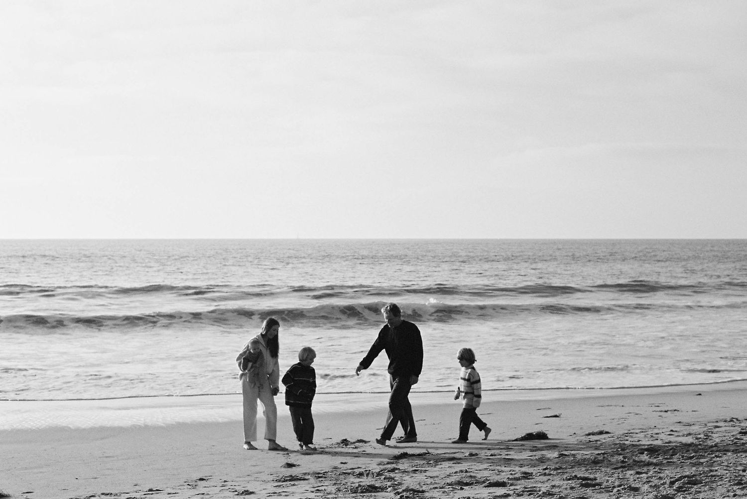 A group of people walk along a misty beach shoreline in black and white as waves roll in from the ocean.