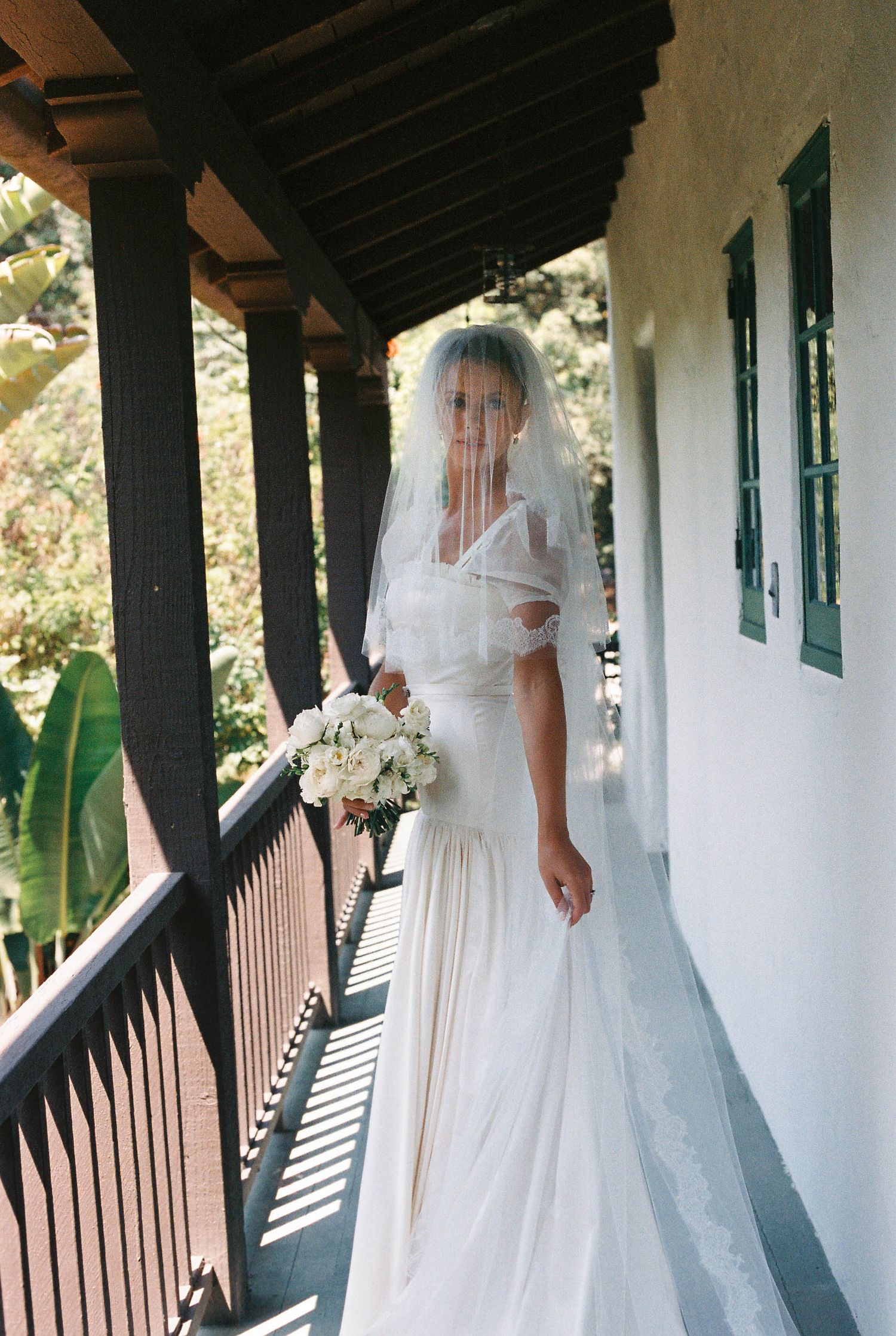 A bride in a flowing white gown and veil poses on a covered porch with white columns and green shutters.