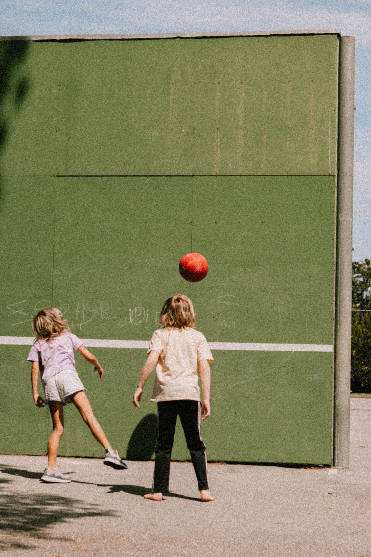 Children playing with a ball against a green wall outdoors.