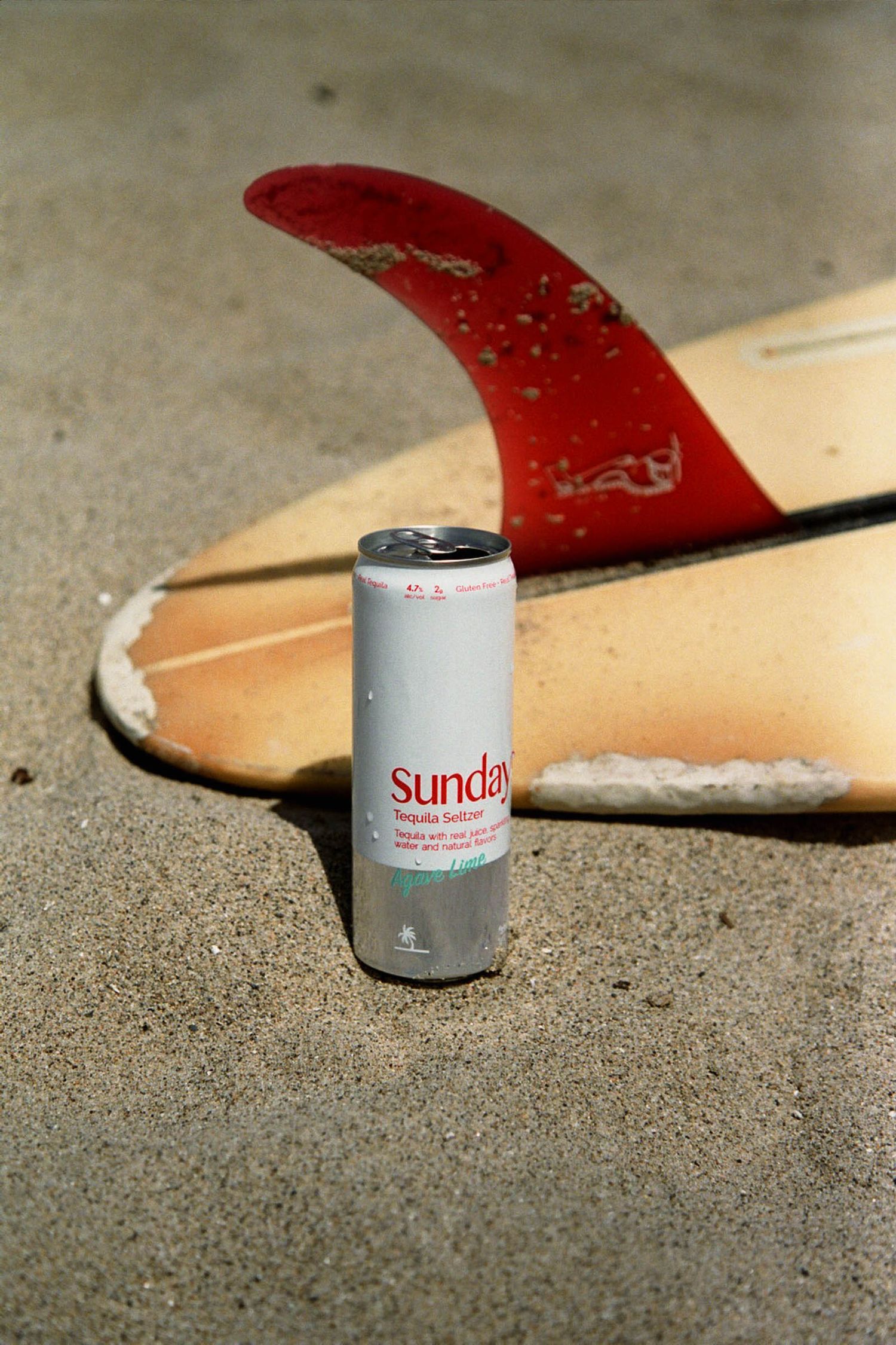A Sundays beverage can sits on sand next to a surfboard with red fin.
