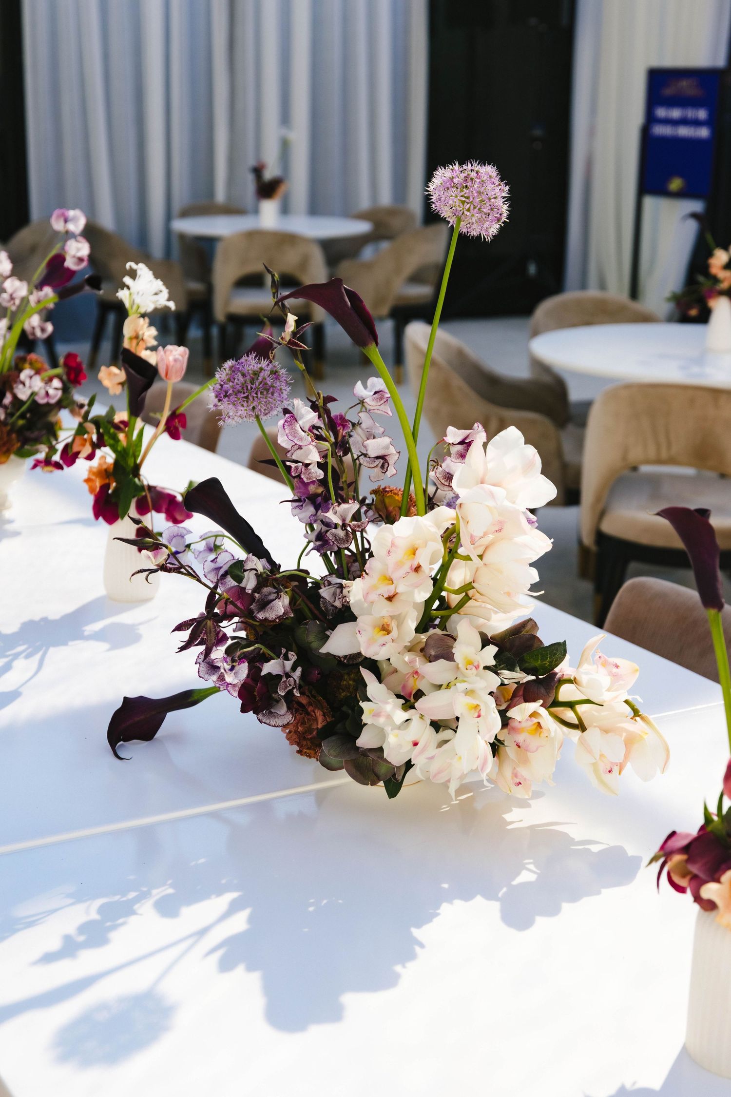 Elegant floral centerpiece with white orchids, purple allium, and dark calla lilies on a white table in a bright venue.