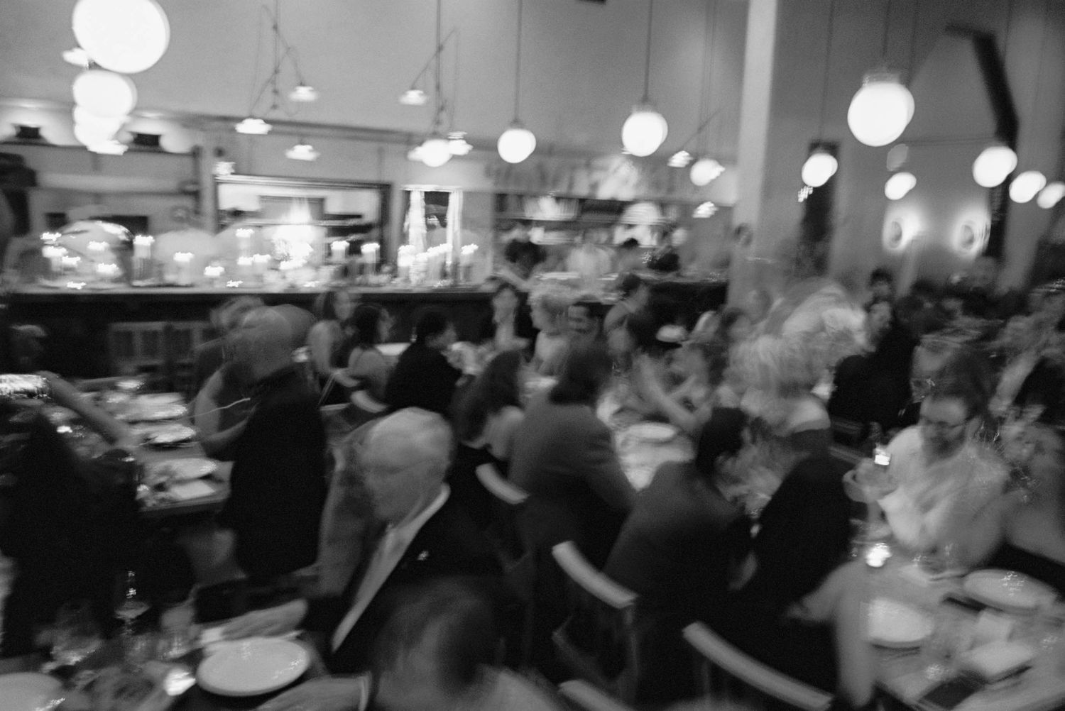 Crowded banquet hall with round tables and white paper lanterns hanging from the ceiling during an evening event.