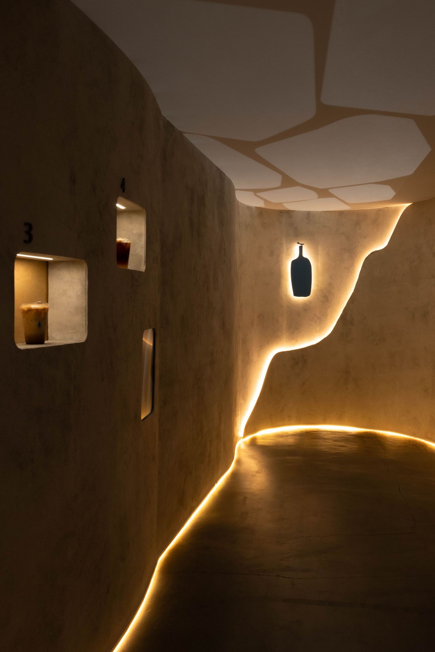 Curved concrete corridor with glowing amber LED strip along an organic edge, numbered wall niches displaying drinks, and a blue bottle logo.