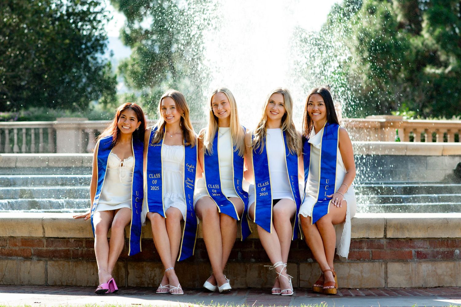 Group of graduates in white dresses and blue stoles sitting together outdoors.