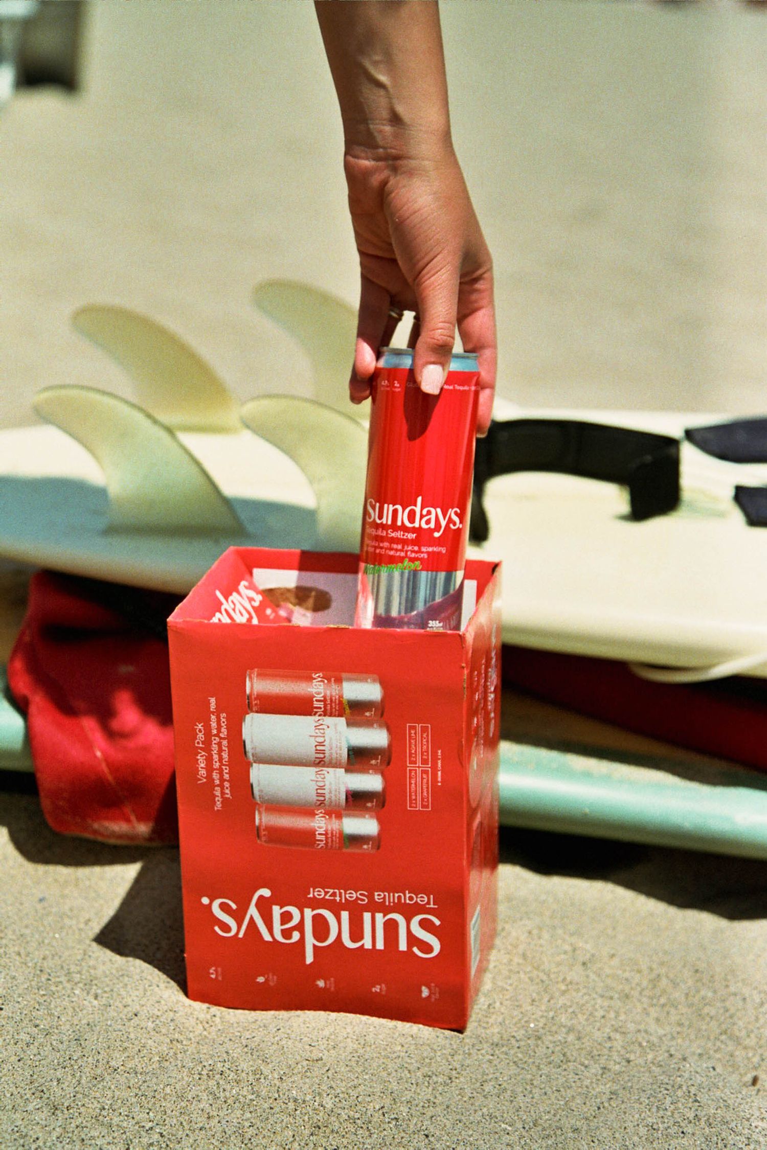 Hand reaching for a Sundays beverage from a red product box near surfboards.