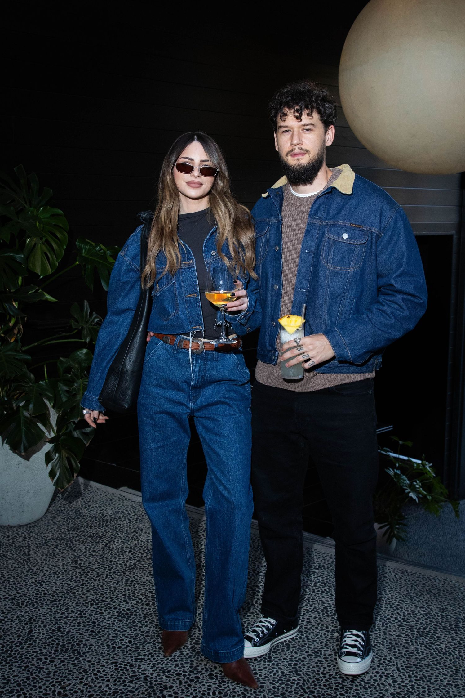 Two people in denim outfits hold cocktail glasses at a dimly lit party.