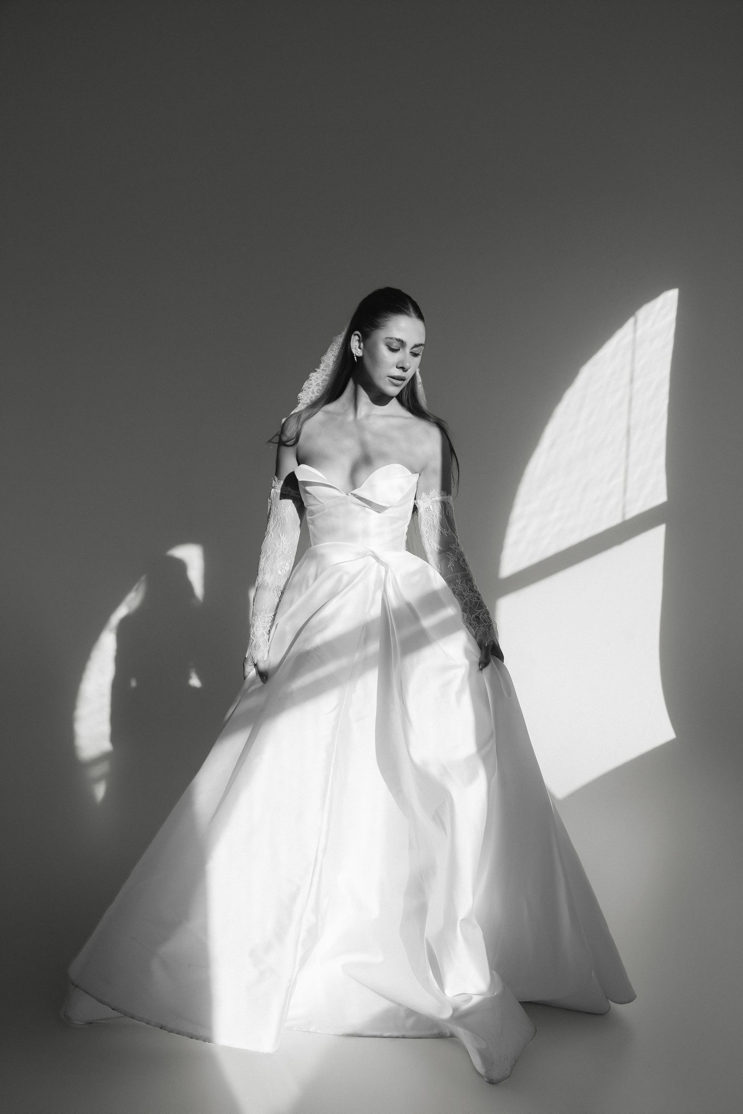 Black and white photo of a bride in an elegant off-shoulder ball gown wedding dress, bathed in dramatic window light.