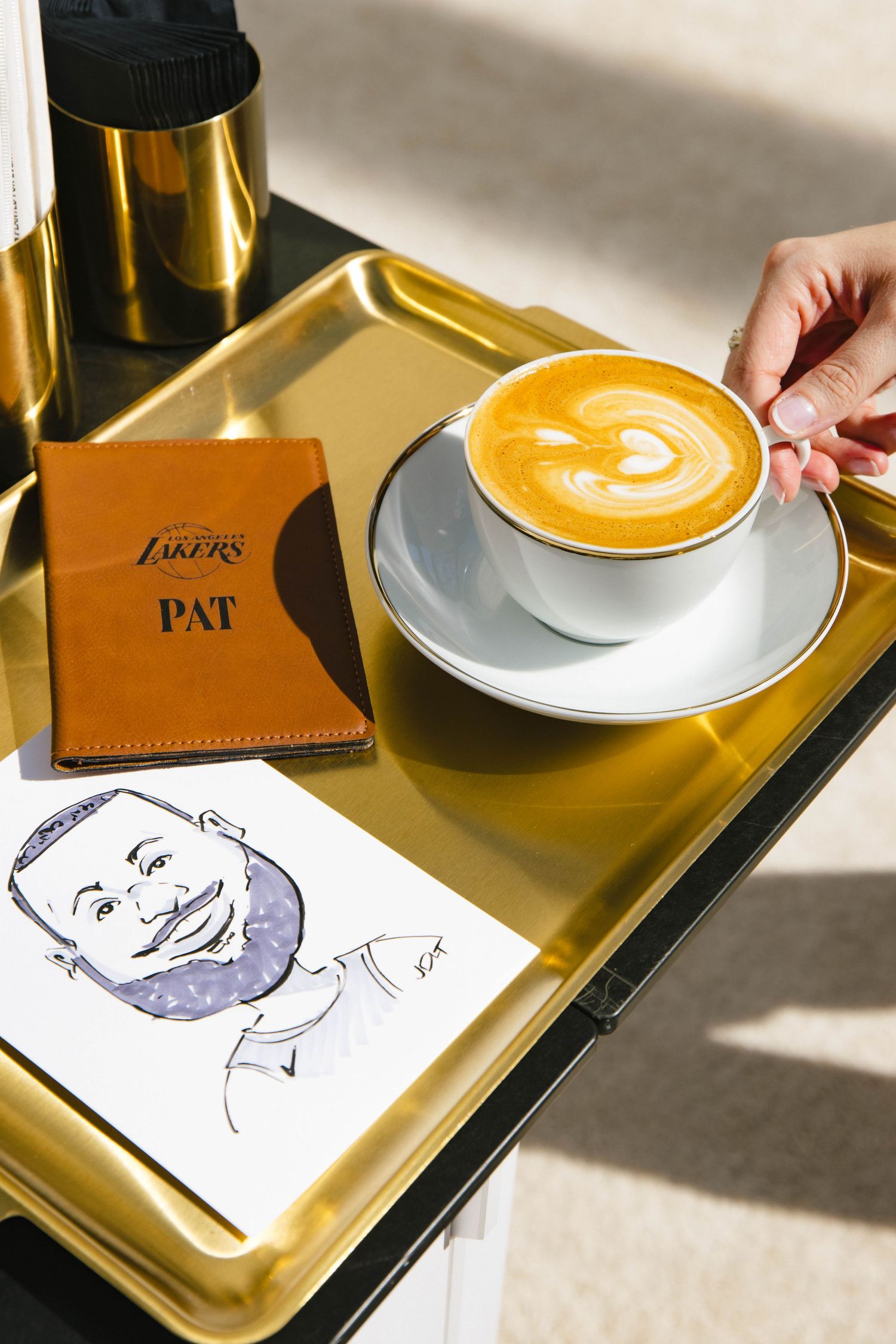 Gold tray with latte art, Lakers branded wallet labeled PAT, and a caricature drawing in sunlight.