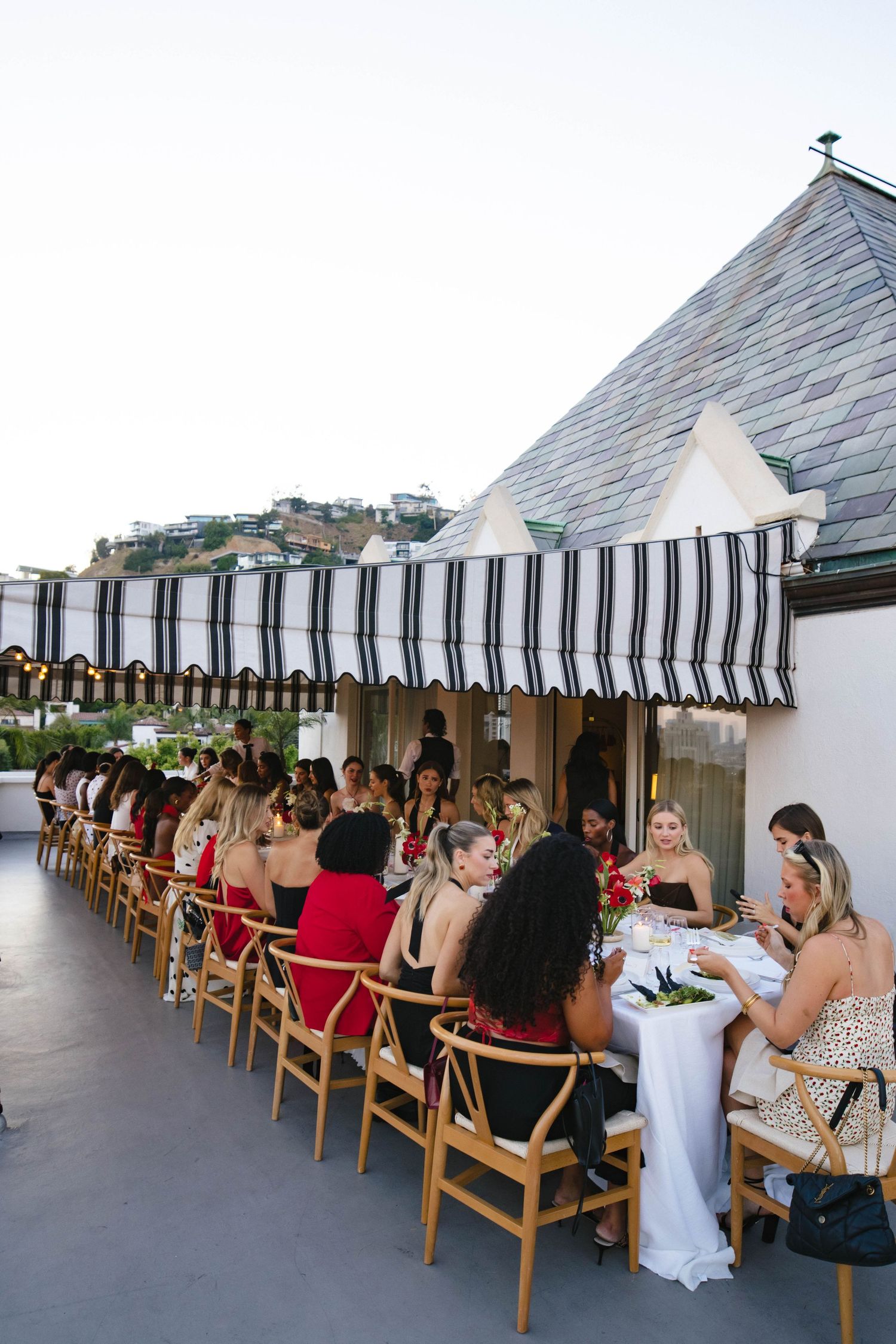 Outdoor rooftop dining event with striped awning and guests seated at long table.