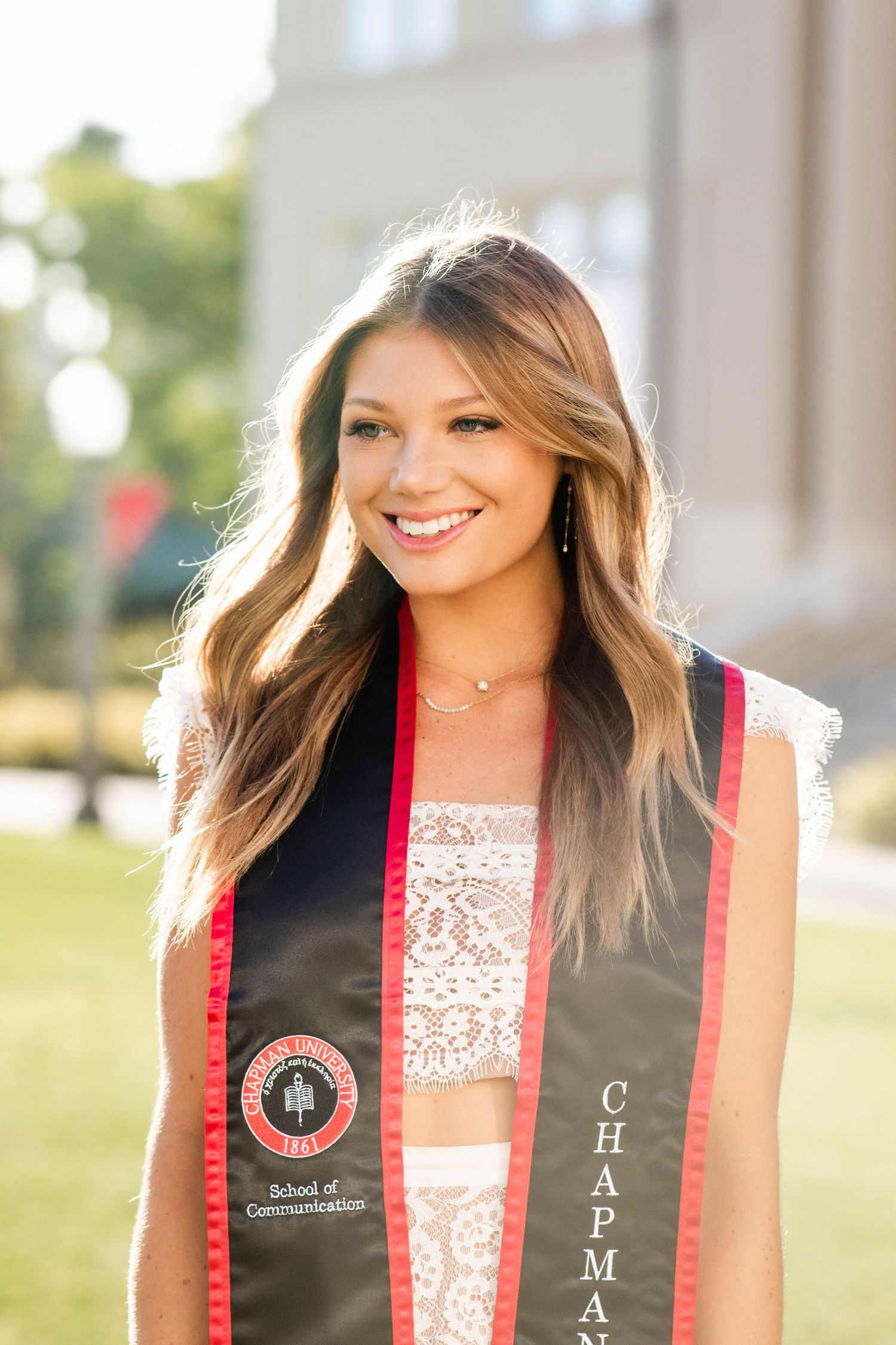 Graduate wearing black and red stole with university insignia smiling outdoors.