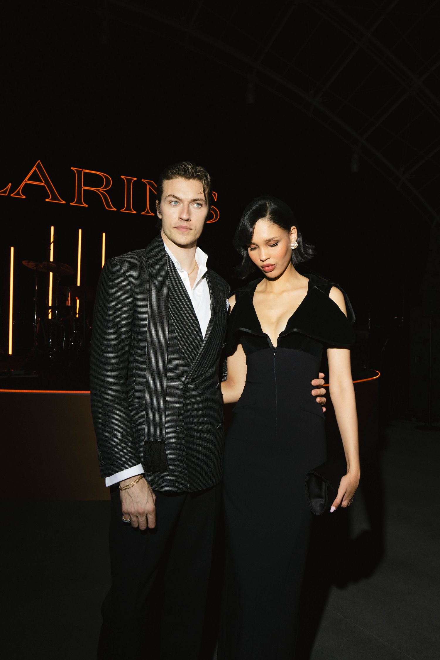 Elegant couple poses at Clarins event, man in pinstripe suit and woman in black off-shoulder gown.