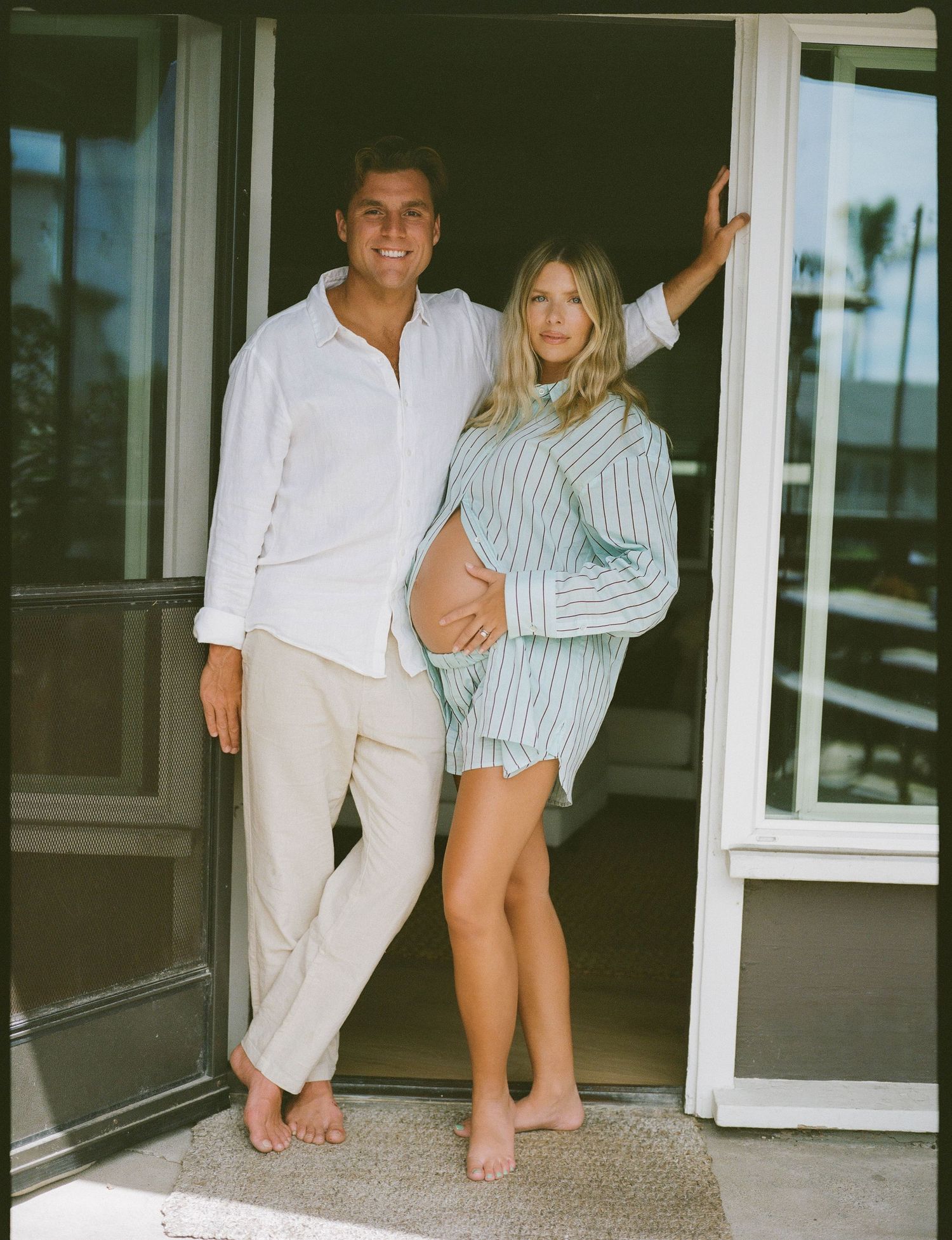 Couple posing in doorway, man in white shirt and woman in striped outfit cradling baby bump, barefoot outdoors.