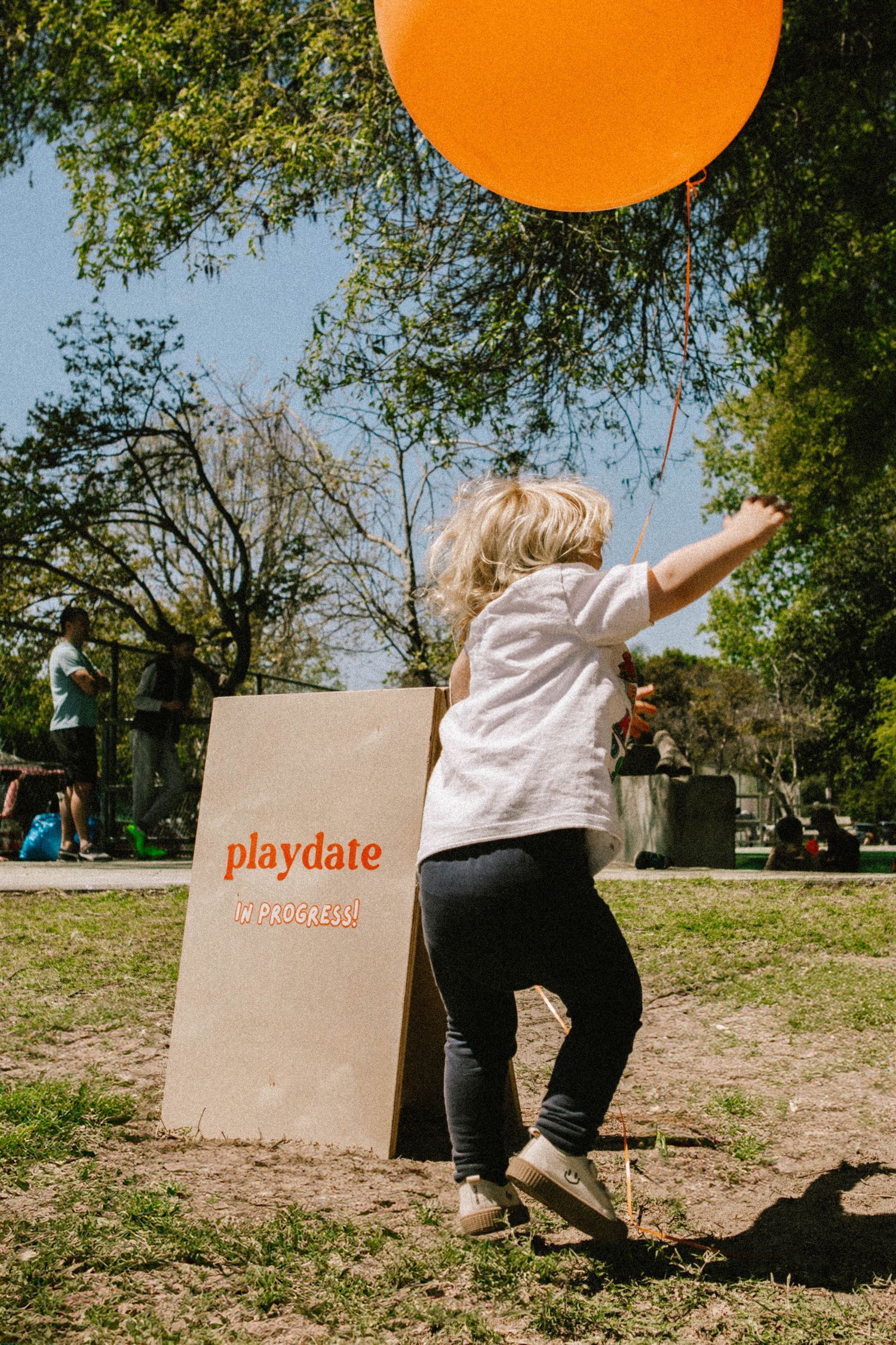 Child jumping to catch an orange balloon next to a Playdate branded box in a sunny park.