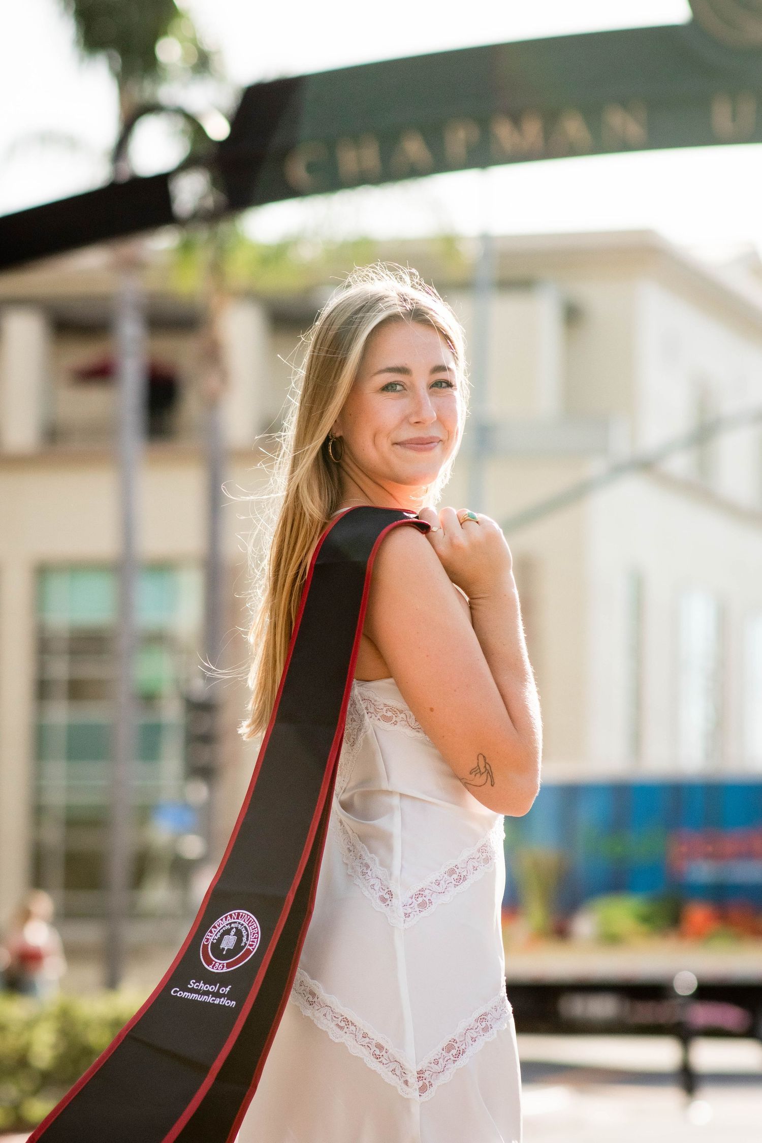 Student in a white dress smiles while carrying a guitar case on campus at Chapman University.