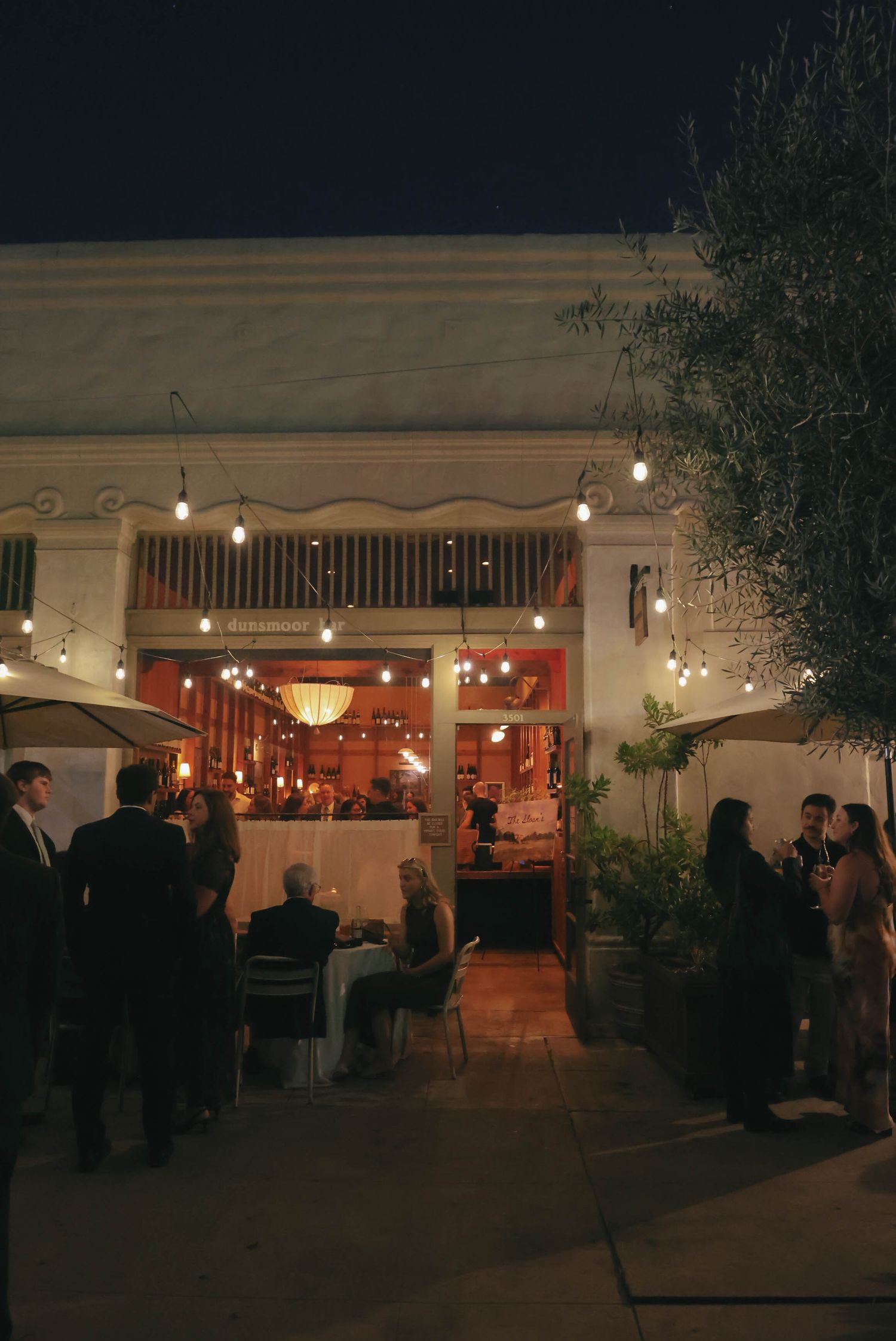 Outdoor restaurant patio at night with warm lighting and guests dining.