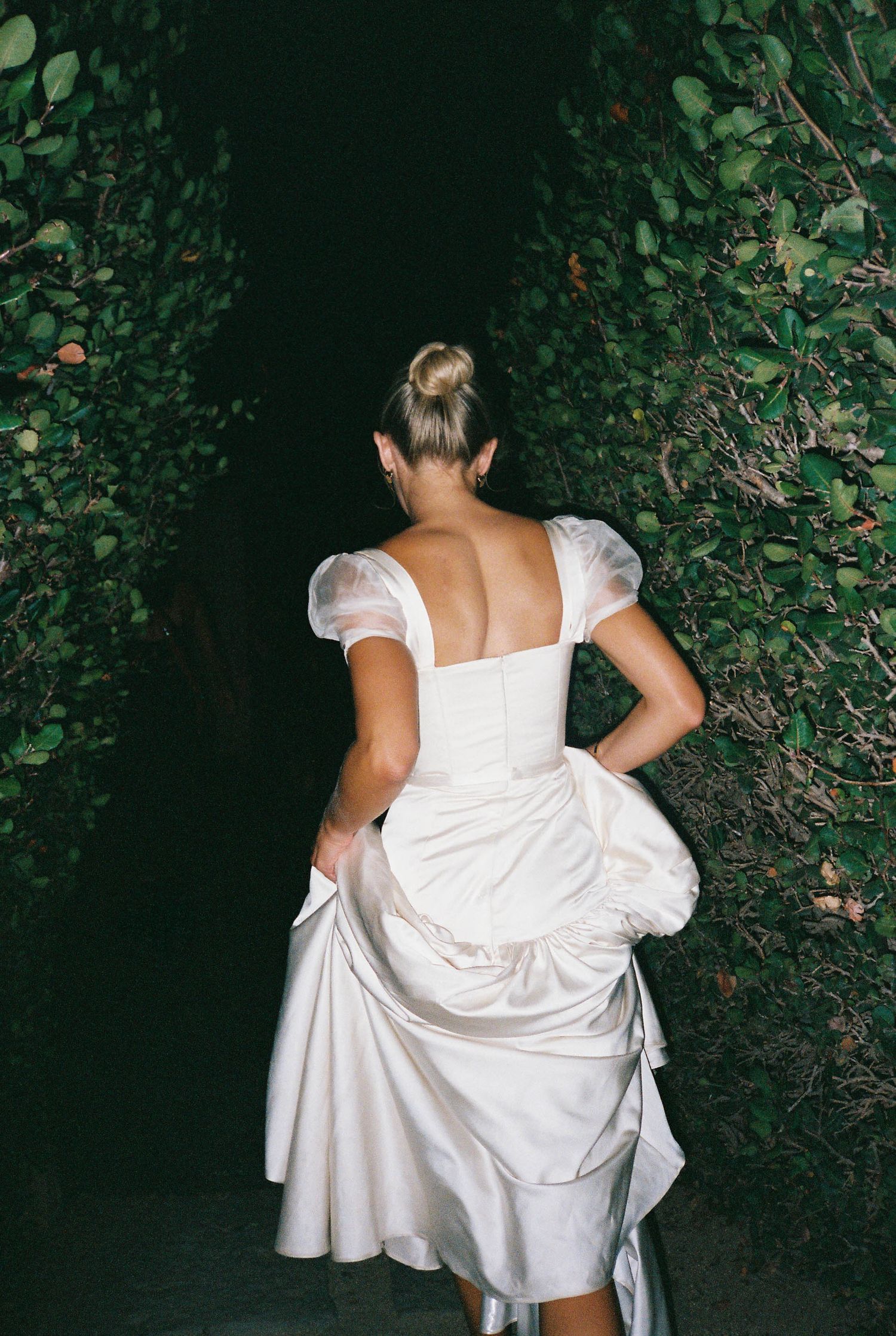 Back view of someone in a white dress walking through a garden with lush greenery.
