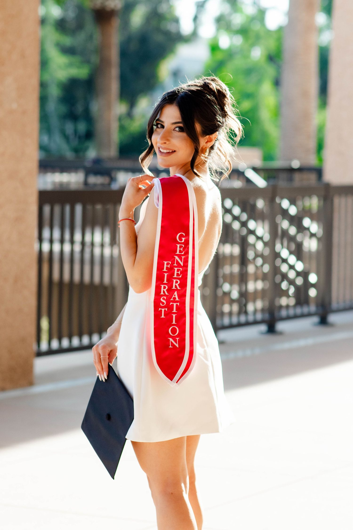 Graduate in white dress poses with graduation cap and red sash on campus.