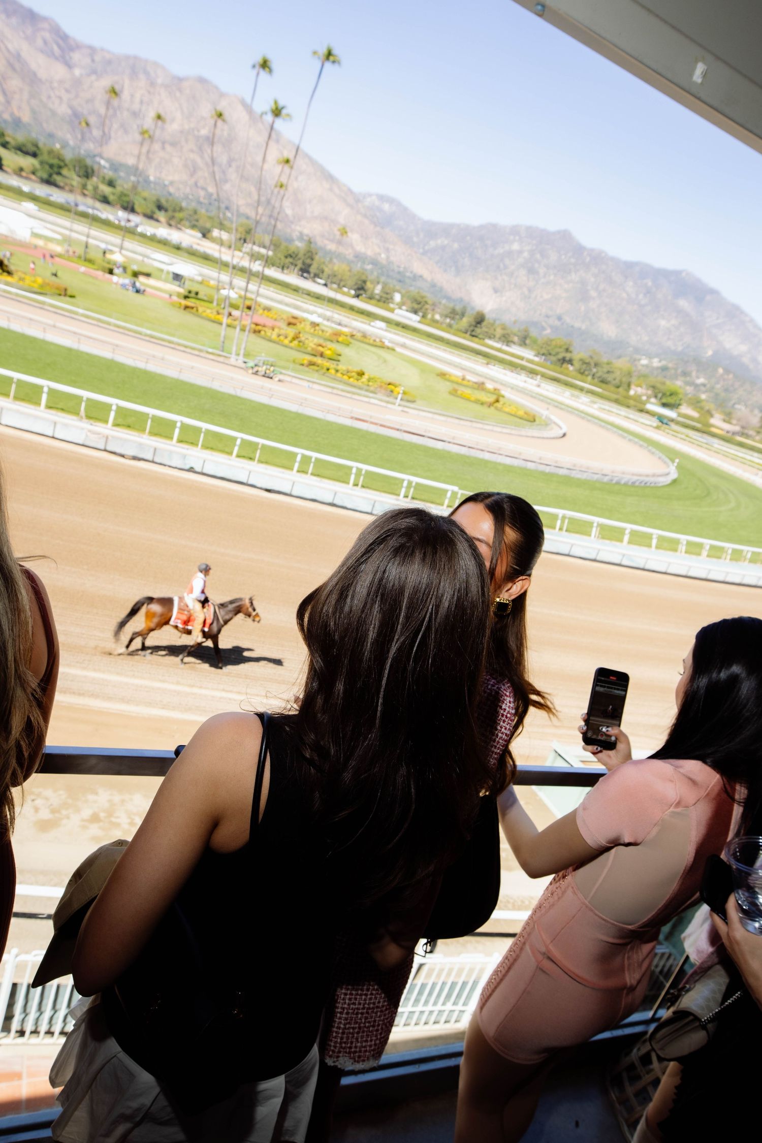 Spectators watch from the viewing area as a horse and rider exercise on the racetrack with mountains in the background.