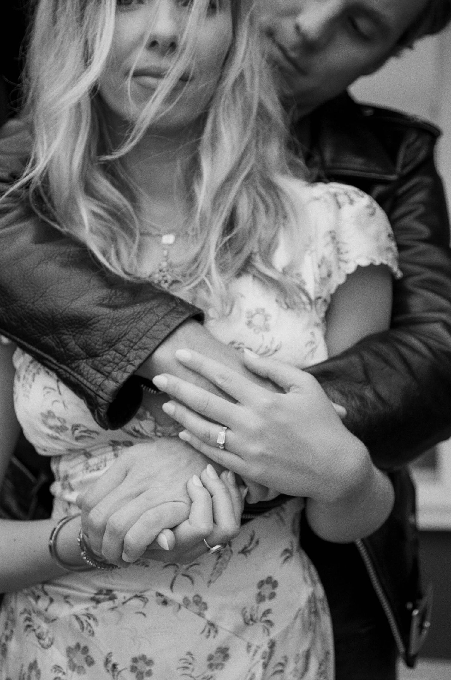 Black and white romantic photo showing couple embracing, with focus on hands and floral dress details.