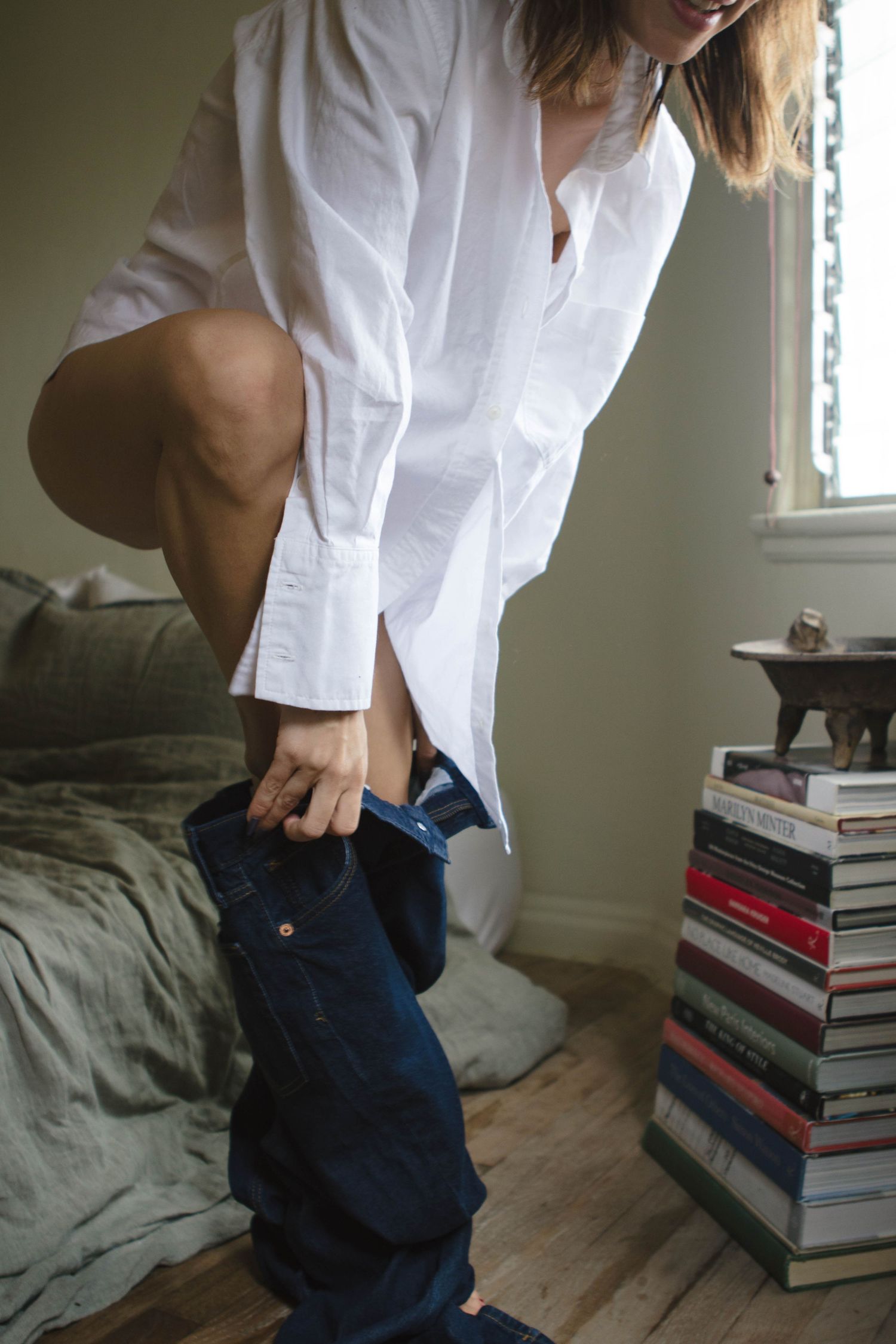 A person in a white button-down shirt and jeans sits on a bed next to a stack of books.