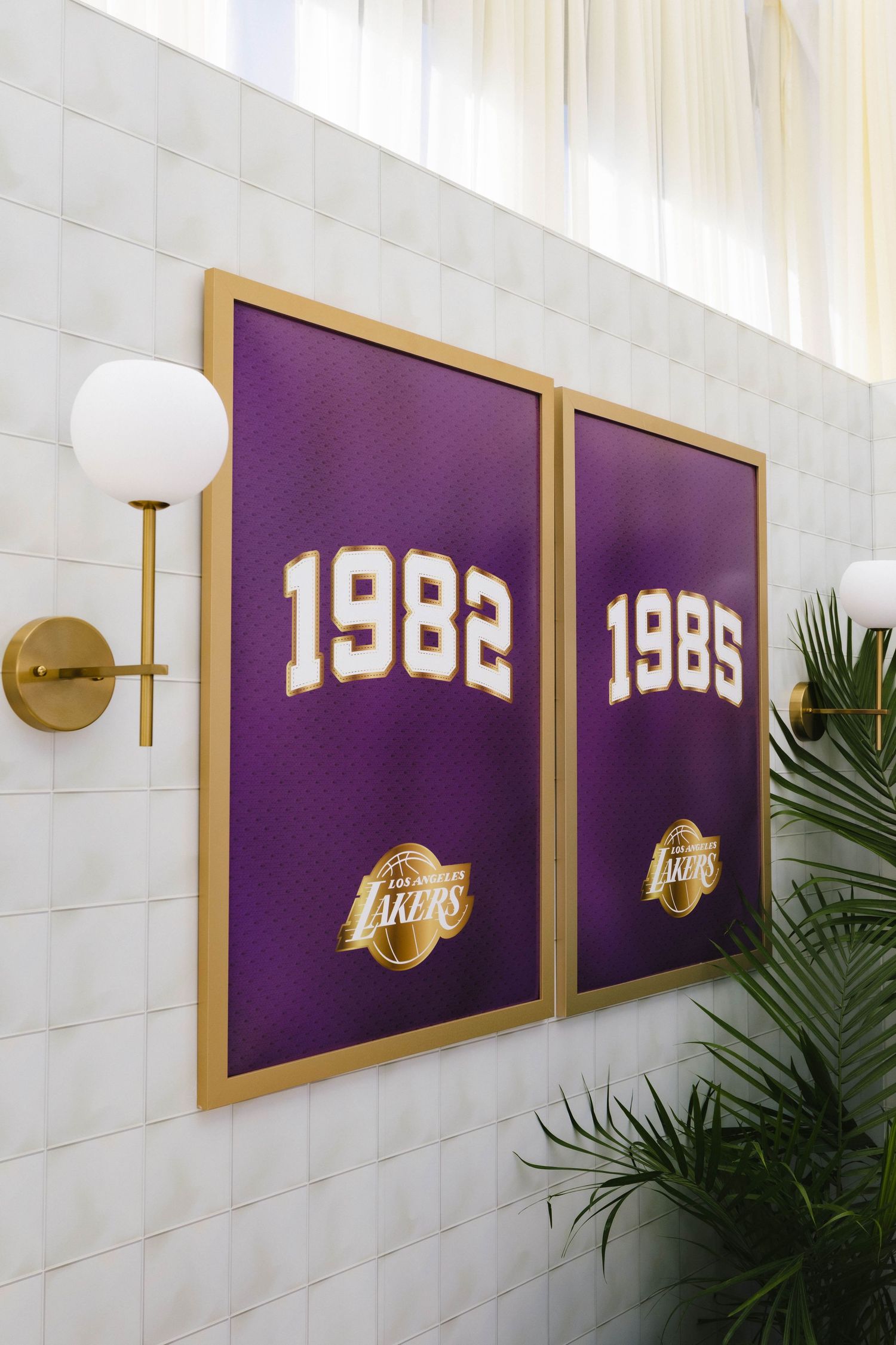 Two gold-framed LA Lakers championship year posters, 1982 and 1985, displayed on white tile wall with brass sconces.