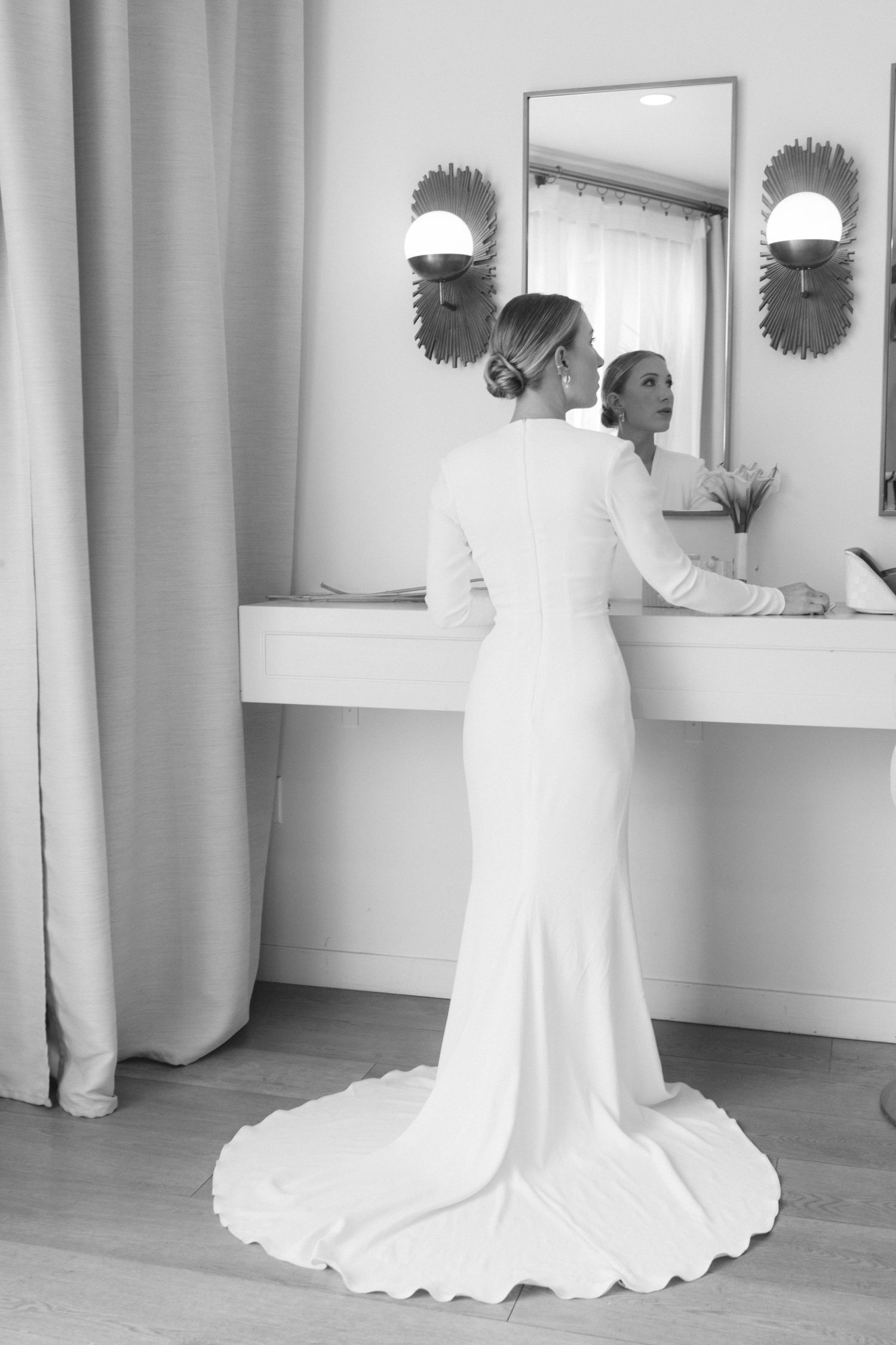 A bride in an elegant white fitted wedding dress with a long train looks at her reflection in a mirror.