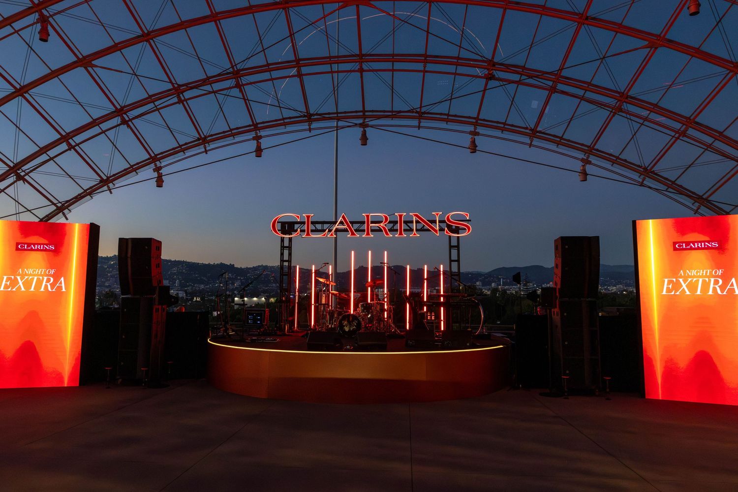 Clarins Extra stage setup at dusk under an arched venue with neon signage and speaker towers.