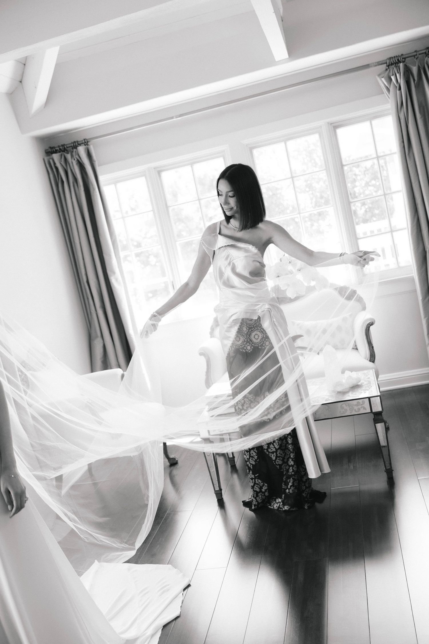 Black and white photo of a bride twirling her sheer wedding veil in a bright, elegant room with hardwood floors.