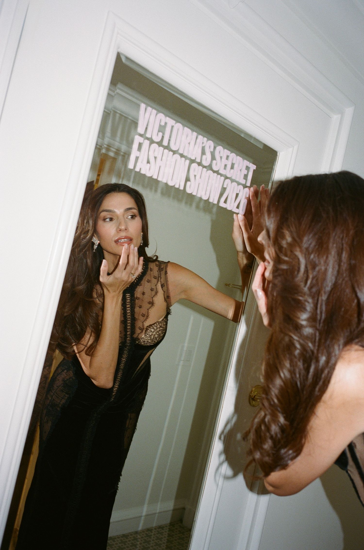 Woman in a black sheer lace dress applies lip color in a door mirror marked Victoria's Secret Fashion Show backstage.